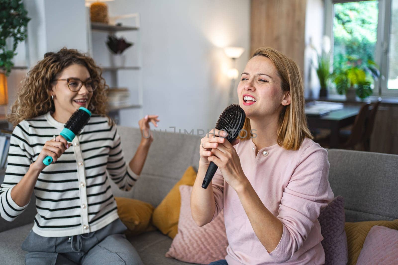 Two friends are sitting on the couch and having a great time singing into hairbrushes instead of microphones.