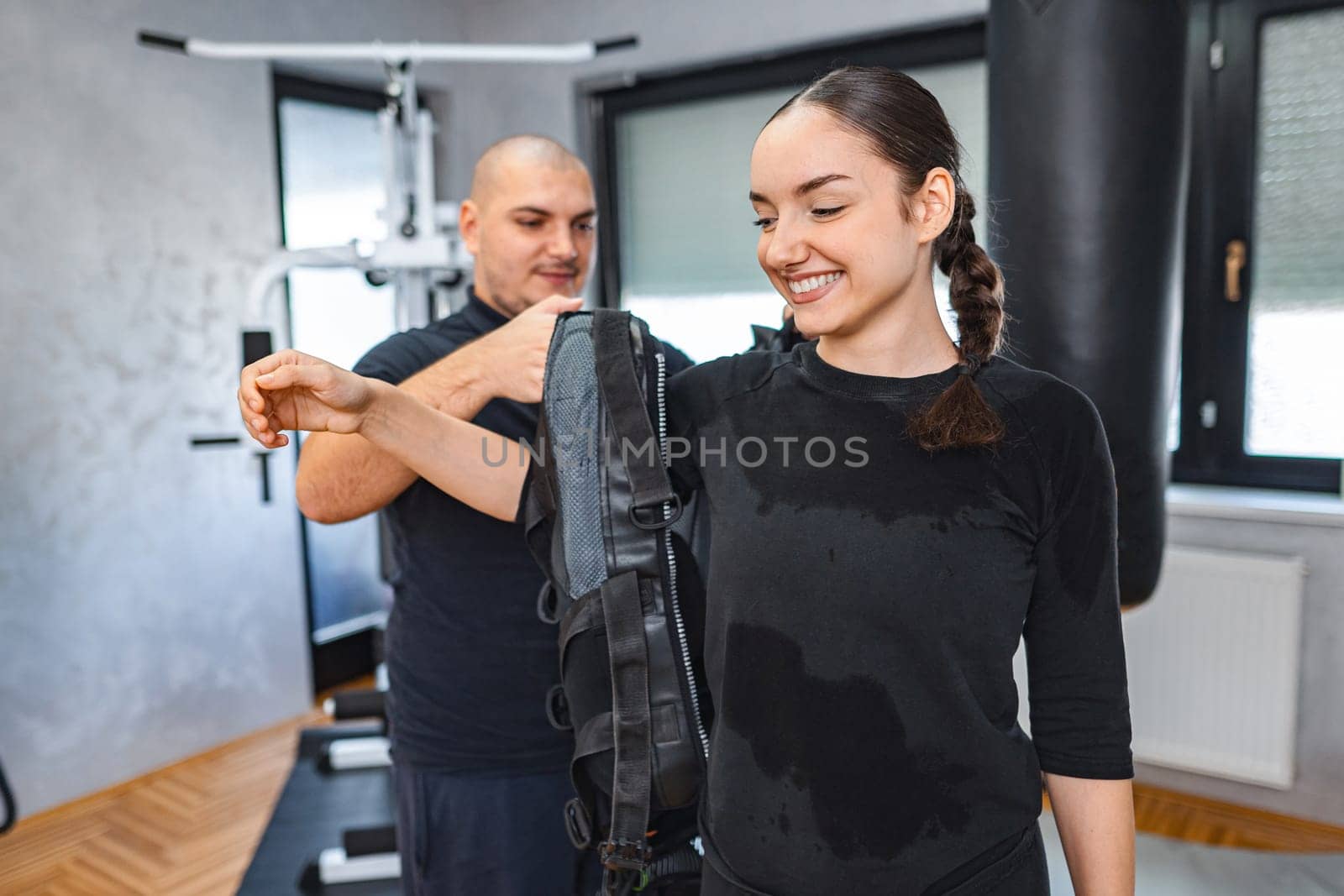 A trainer helps a woman prepare for an EMS workout session in a gym.