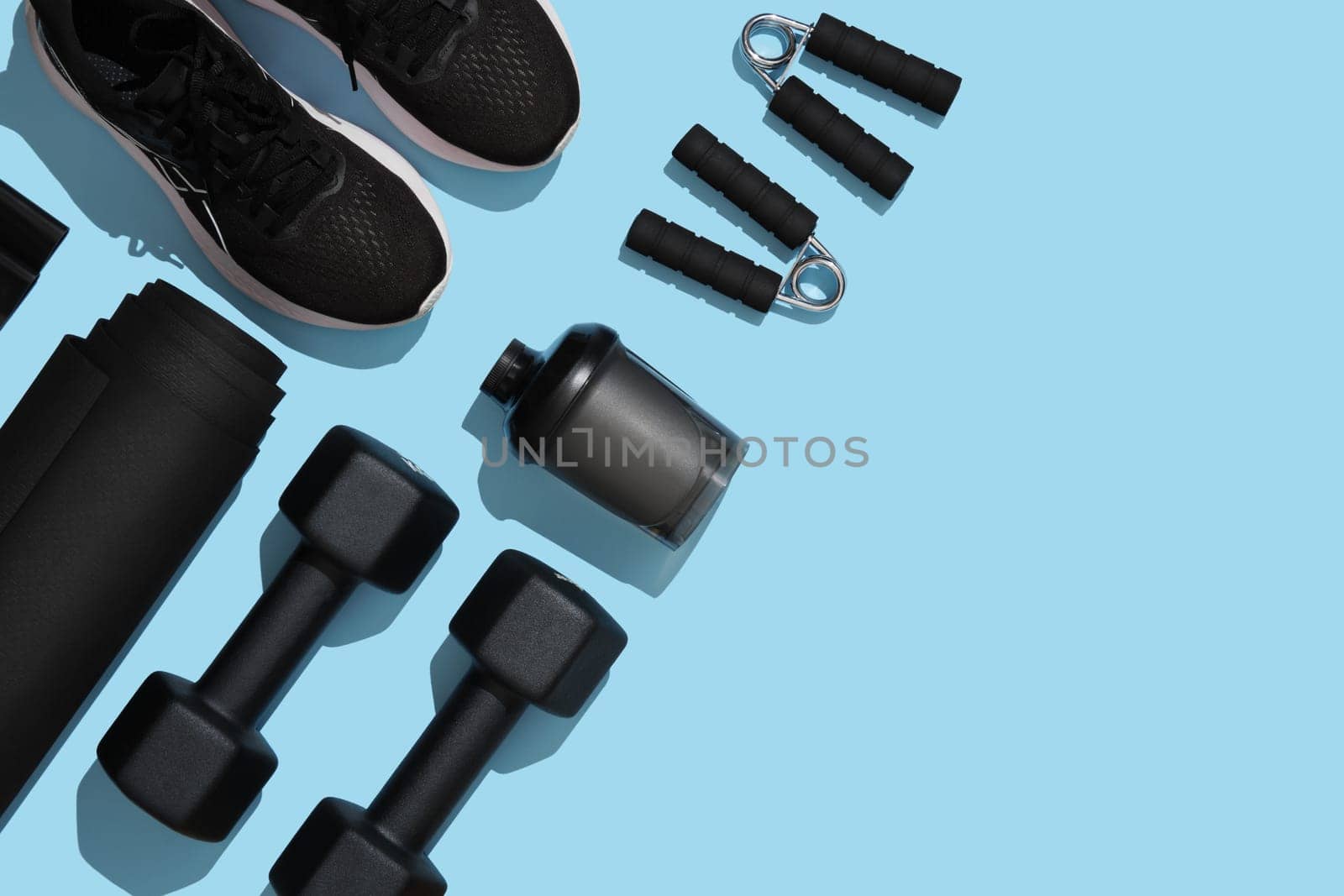 Black workout equipment, dumbbells, mat, hand grips, sneakers and water bottle on blue background. Fitness, sports items, healthy lifestyle concept. Place for your text