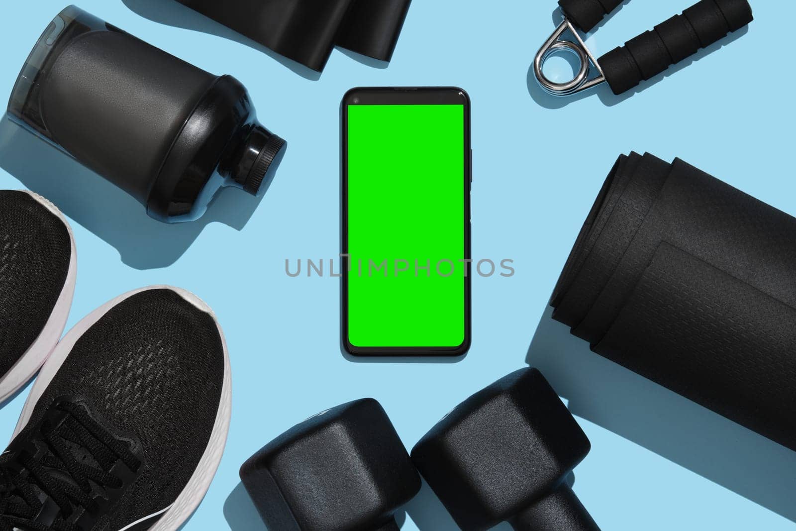 Smartphone mockup with green screen on the background of black fitness items, dumbbells, mat, sneakers, water bottle, sports elastic band. Demonstrating copy space for mobile application