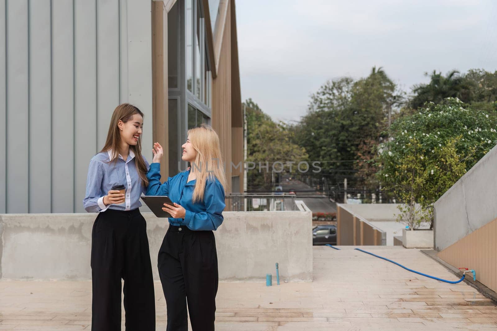 Two diverse women engaging in a lively conversation while holding a tablet and coffee outdoors in a contemporary setting.