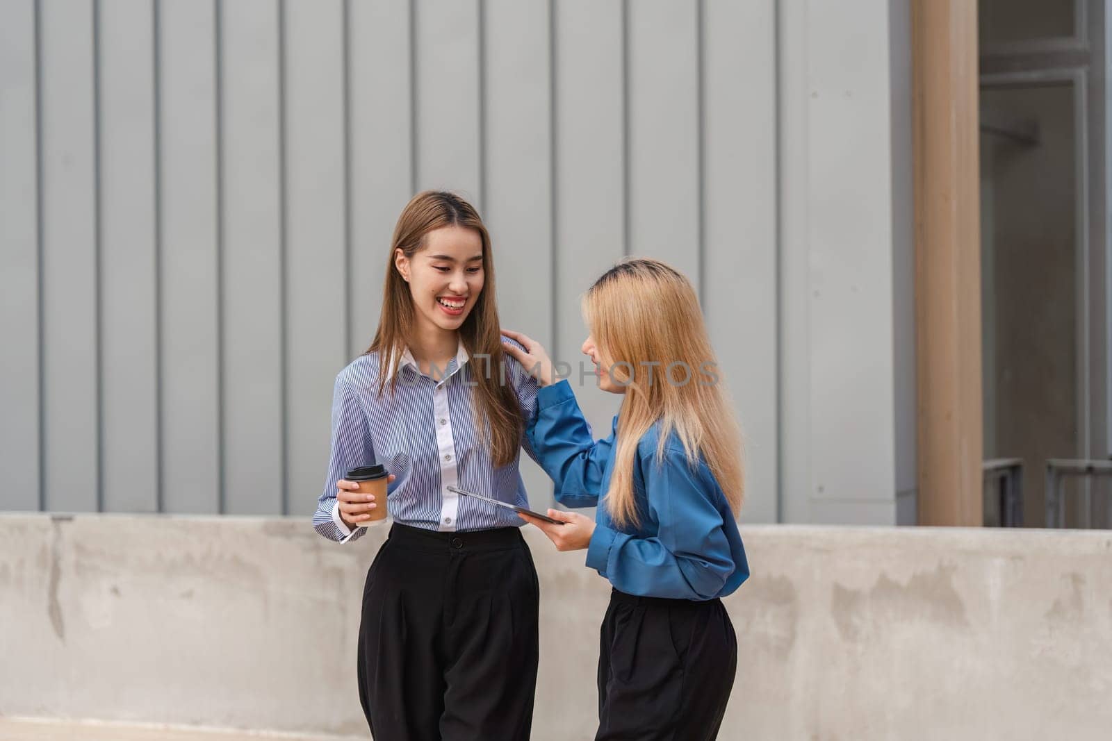Two diverse women walking side by side and discussing ideas, with one holding a coffee cup and the other a tablet, in a modern outdoor setting.