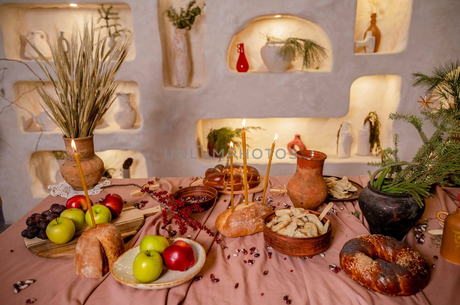 table set for Christmas or another holiday. candles, apples, bread, berries, grapes are burning on the table. in the background is a decorative wall with lighting and clay pots.