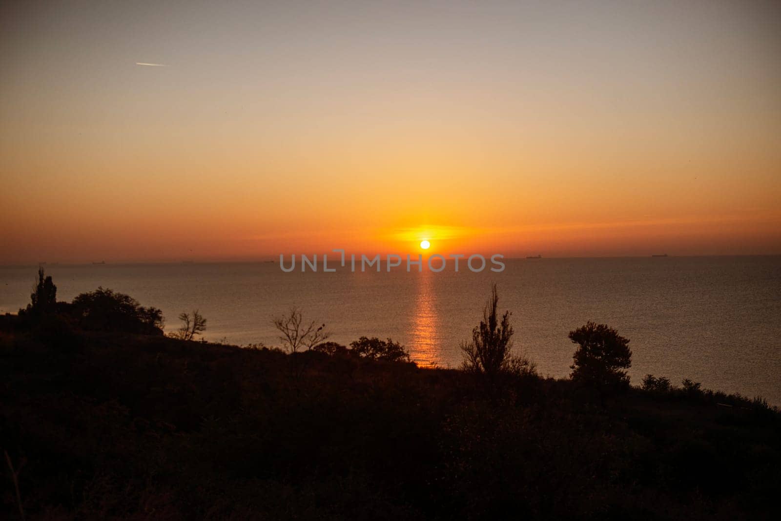 The bright sun rises over the sea. A ray of sun creates a path of warm light on the sea surface. The sea is calm, a ship is visible on the sea horizon, rocks are in the foreground,sun in the center