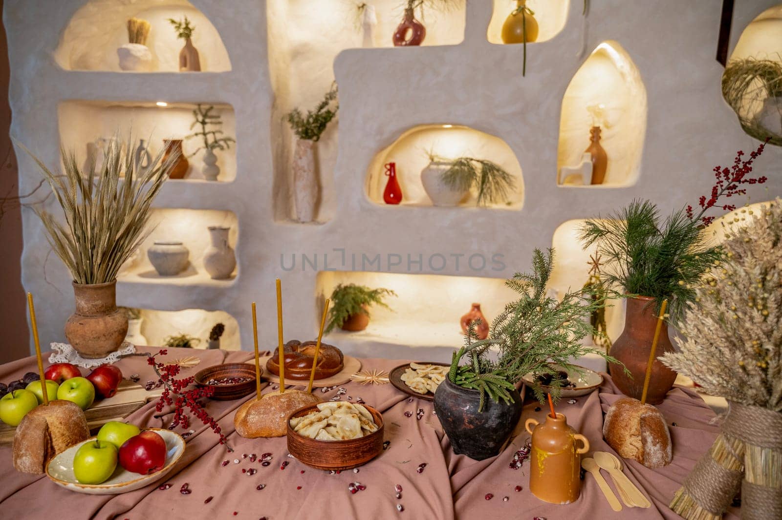 table set for Christmas or another holiday. candles, apples, bread, berries, grapes, dumplings are burning on the table. in the background is a decorative wall with lighting and clay pots. horizontal