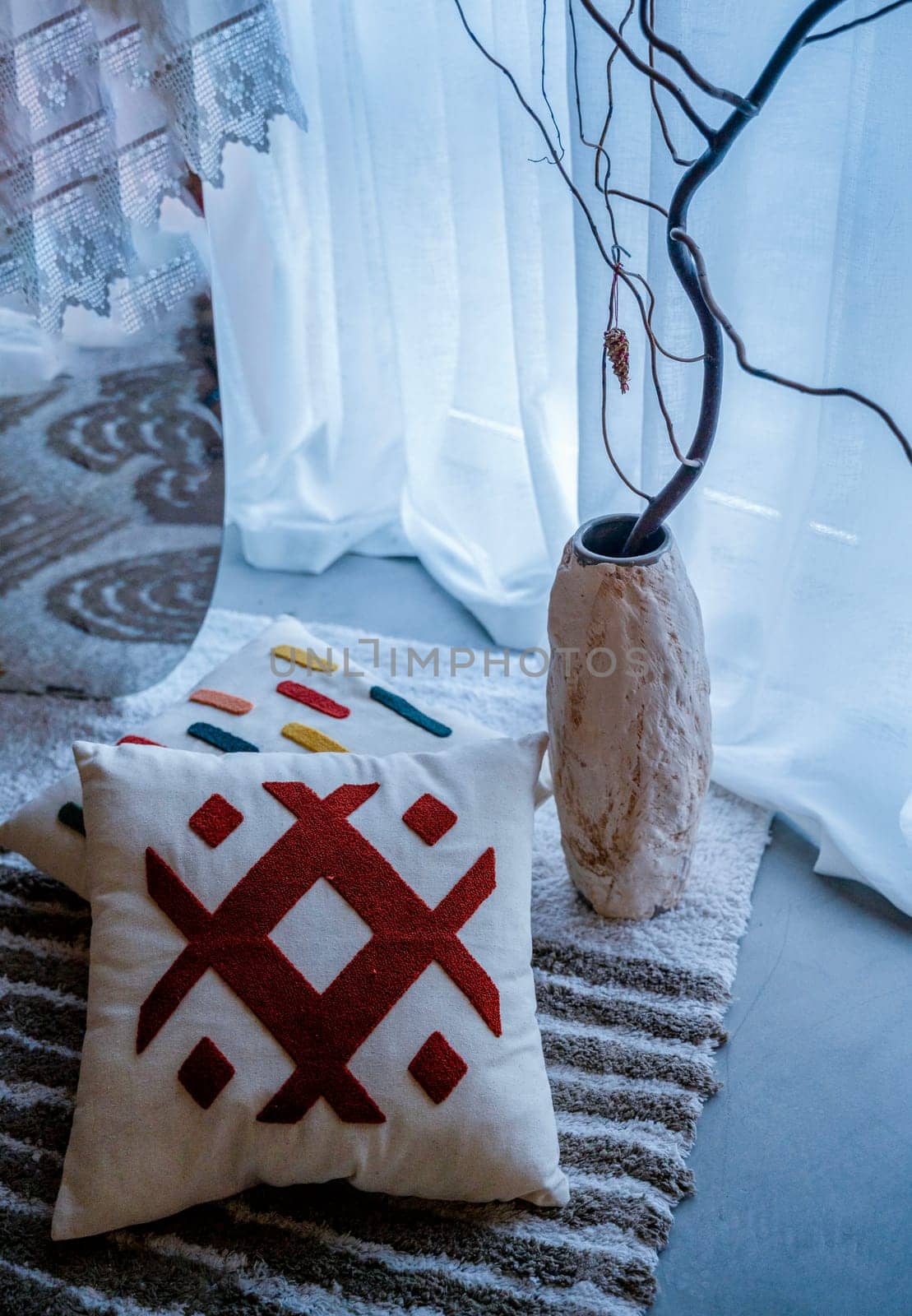 Two decorative pillows lie on the floor next to a light decorative vase. There is also a striped rug on the floor. Light from a window covered with a curtain.