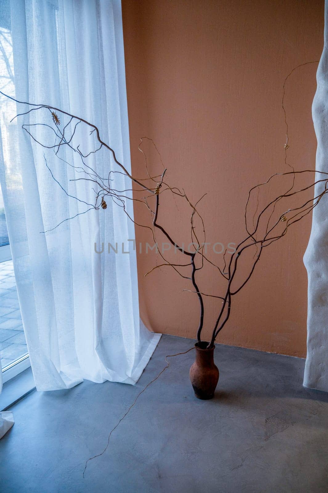 On the gray concrete floor there is a clay vase with a decorative branch. Against the background of a beige saturated wall. Light from the window. The window is very large, closed with a curtain
