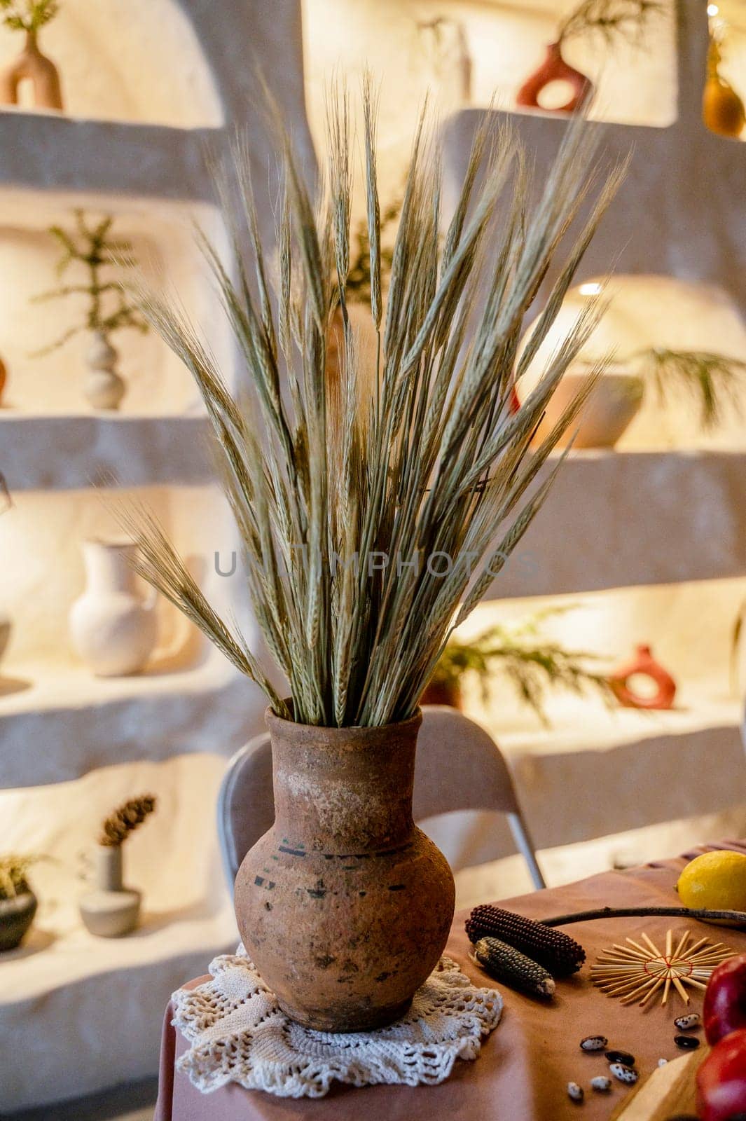 Still life with an antique clay pot and ears of wheat. The vase is of a pleasant brown color. The photo is in warm shades, behind is a backlit wall. There is a white handmade napkin on the table