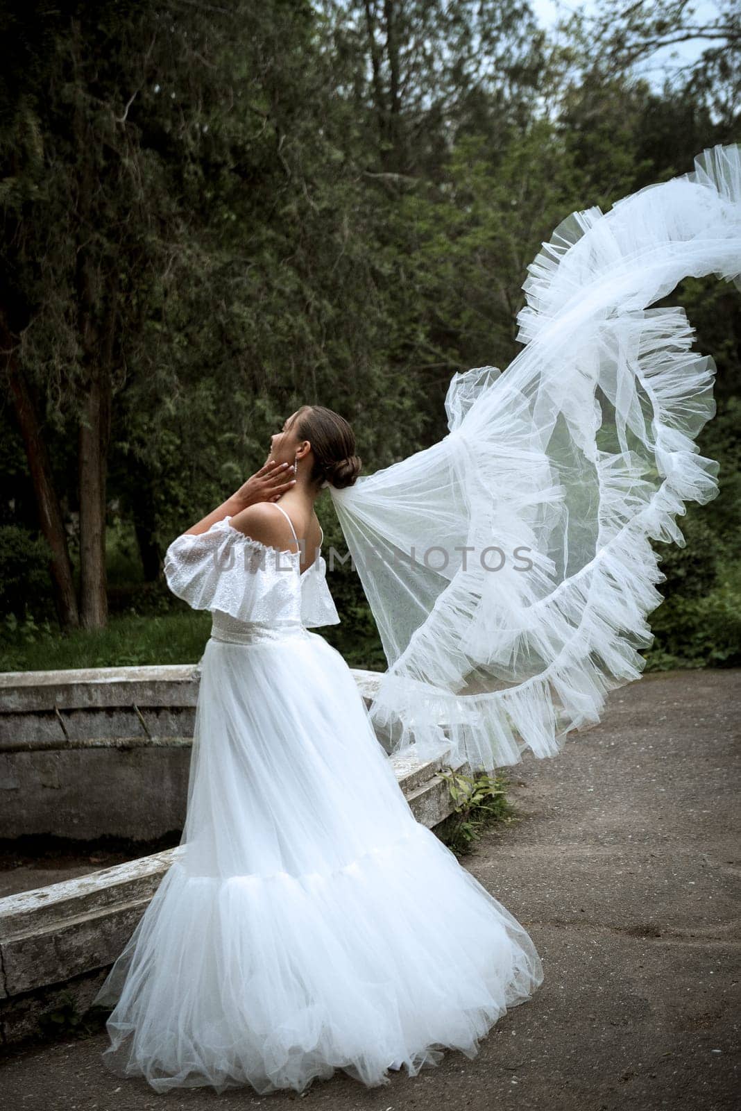 beautiful bride with long veil against the background of trees and old fountain. brunette girl in long white fluffy dress smiles and rejoices. veil flutters in the wind bride looks to the side