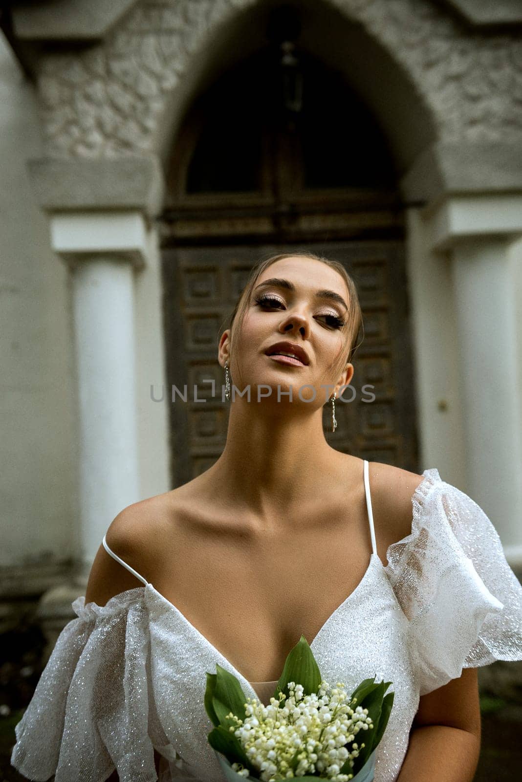 close-up of bride in white dress and with lily of the valley bouquet. dress with open shoulders. behind is old church of 18th century. girl is looking at camera