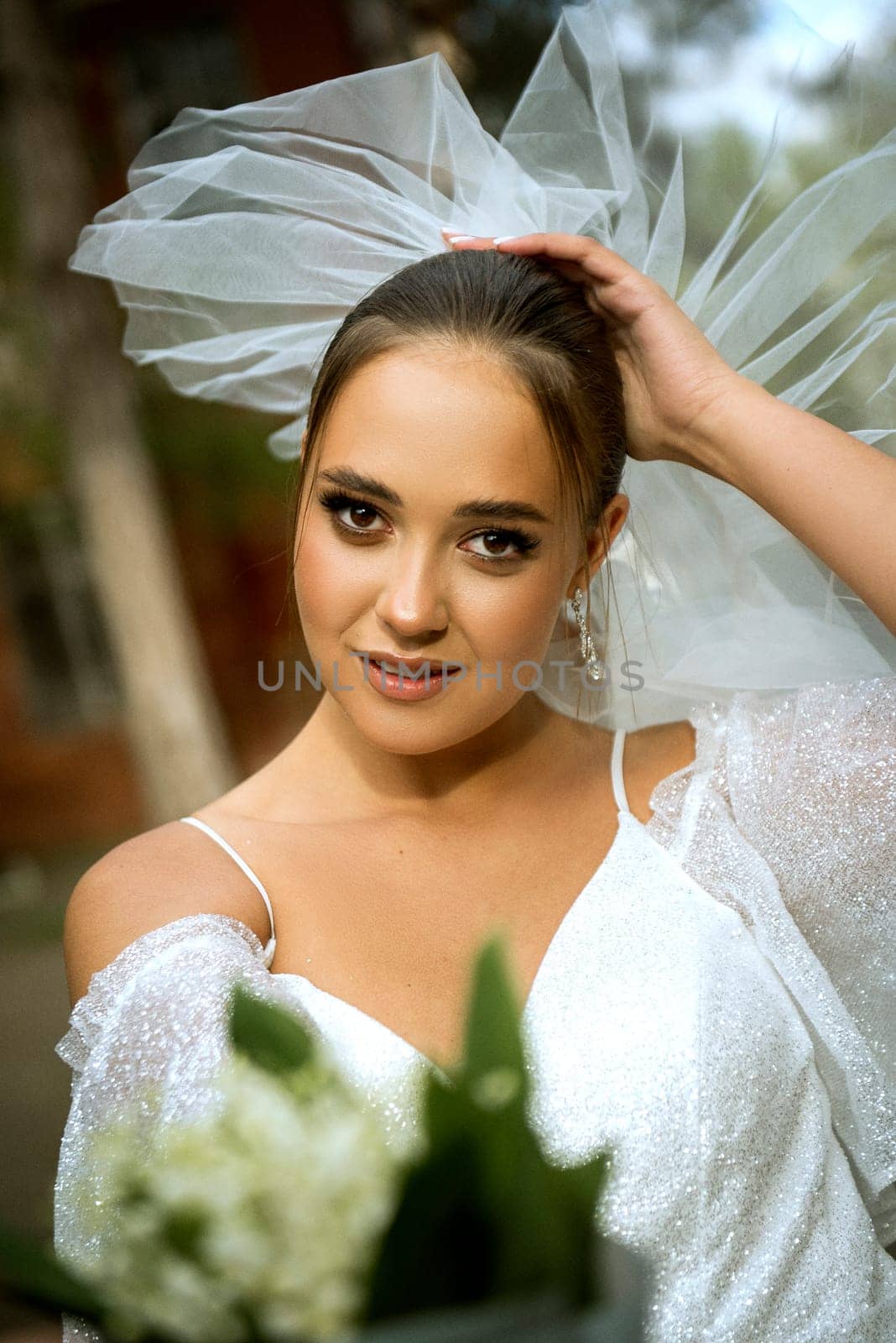 bride in a white dress with open shoulders, with a veil, a bouquet of lilies of the valley. the brunette has a smooth hairstyle gathered in a bun. she is looking at the camera,