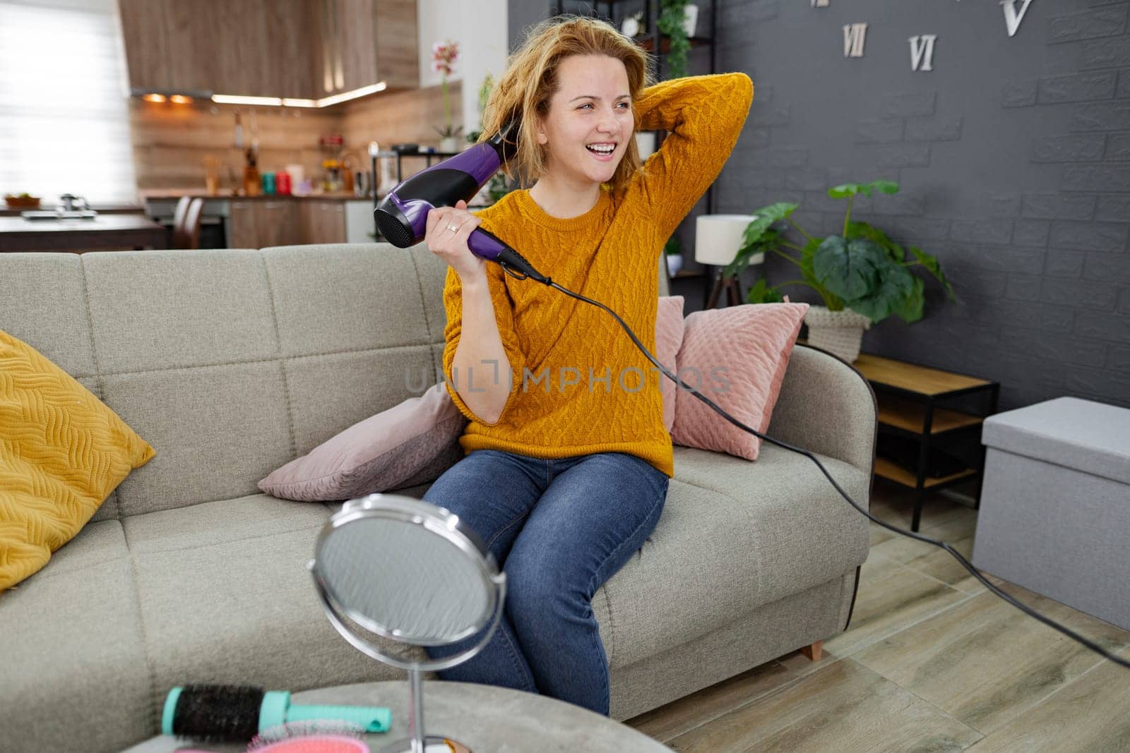 A woman is seated on a couch, using a blow dryer to dry her hair. She is focused on her task, with the blow dryer in hand and hairbrush nearby.