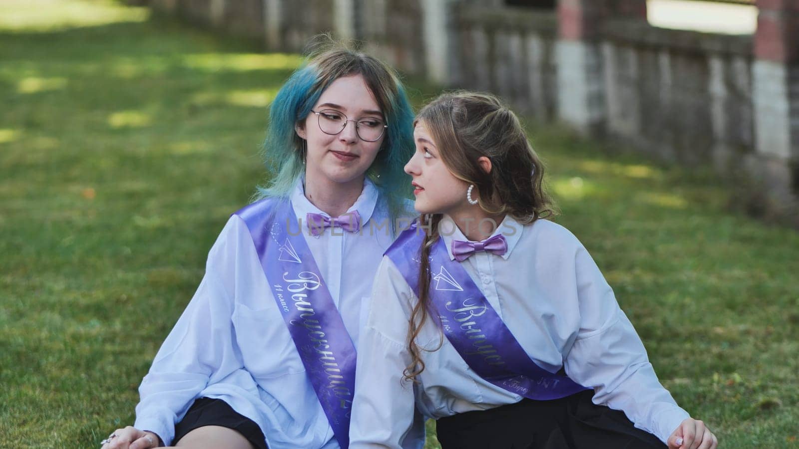 Two joyful graduates in sashes relax on the grass and enjoy a conversation on their final day of school
