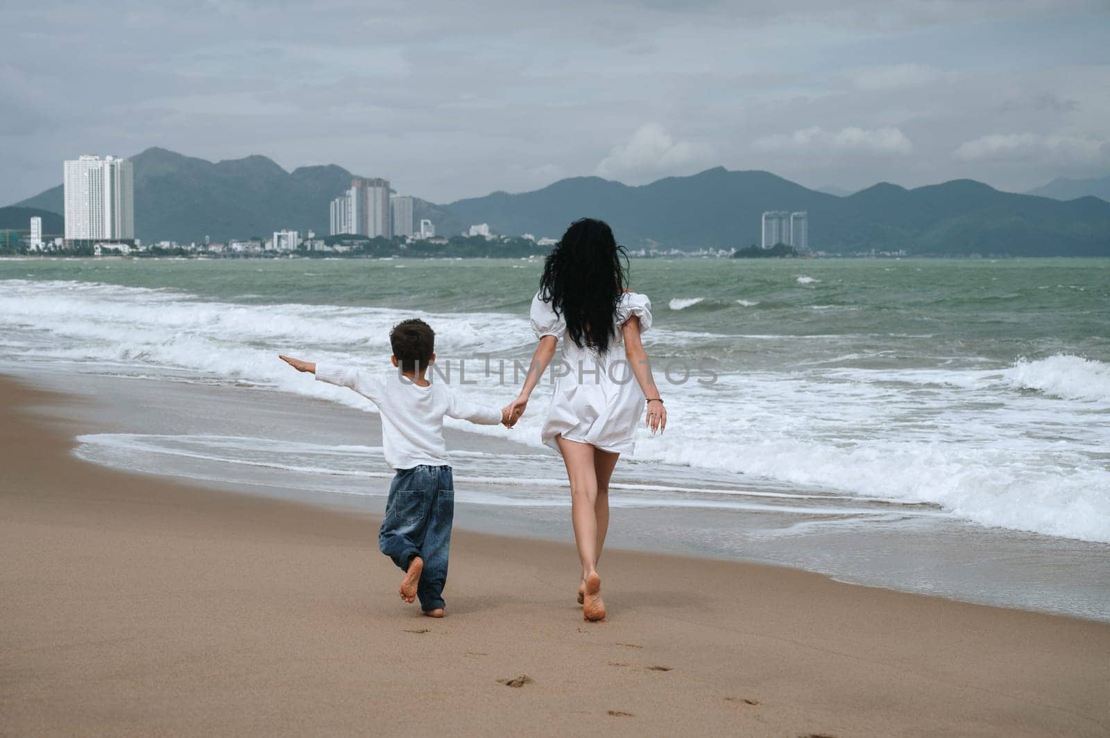 back of happy mom and son walking along the beach by the sea on summer vacation