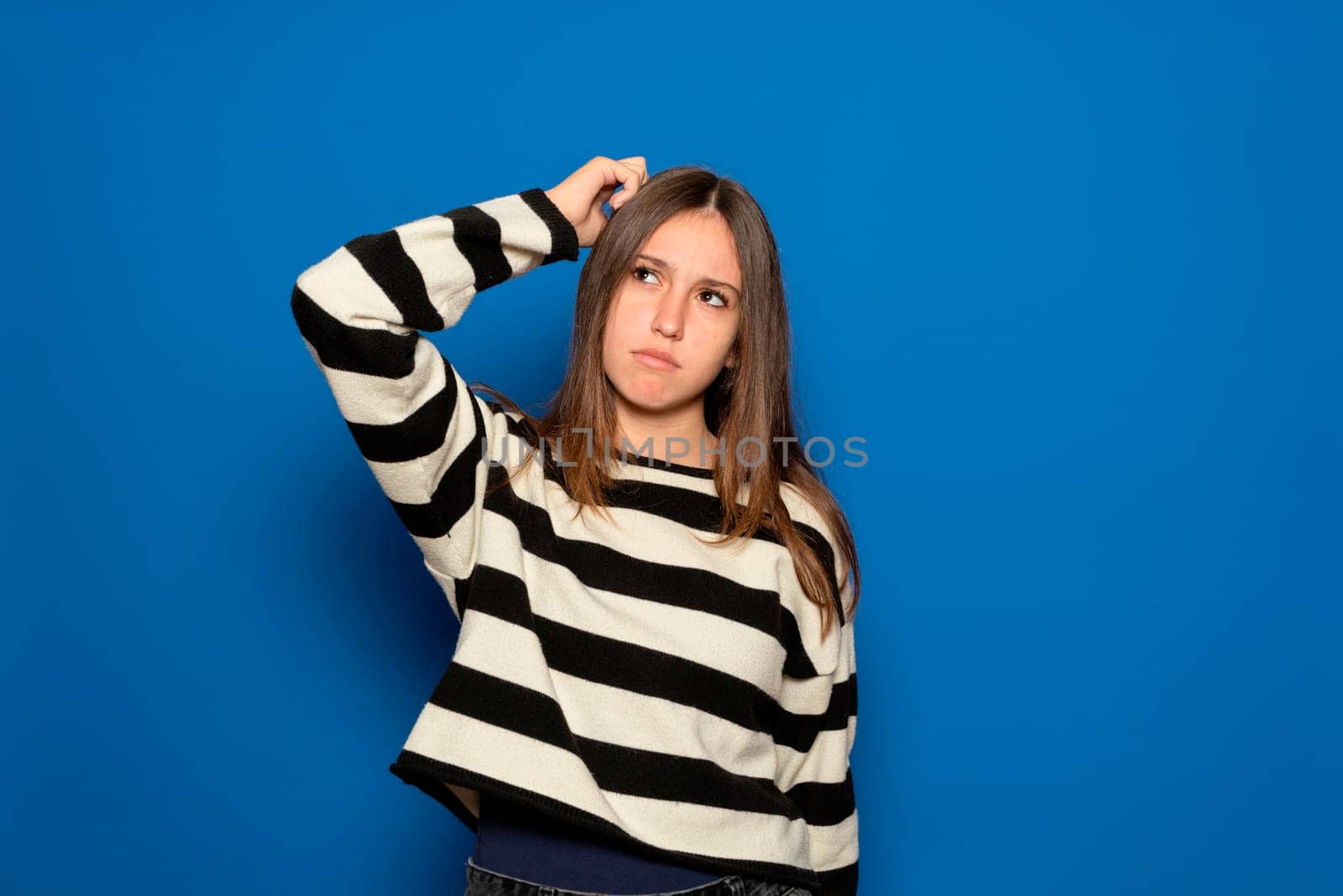 Hispanic teenage girl about 15 years old wearing a striped sweater and a puzzled facial expression, scratches her head in frustration, isolated over a blue background. Hmm, I don't remember.
