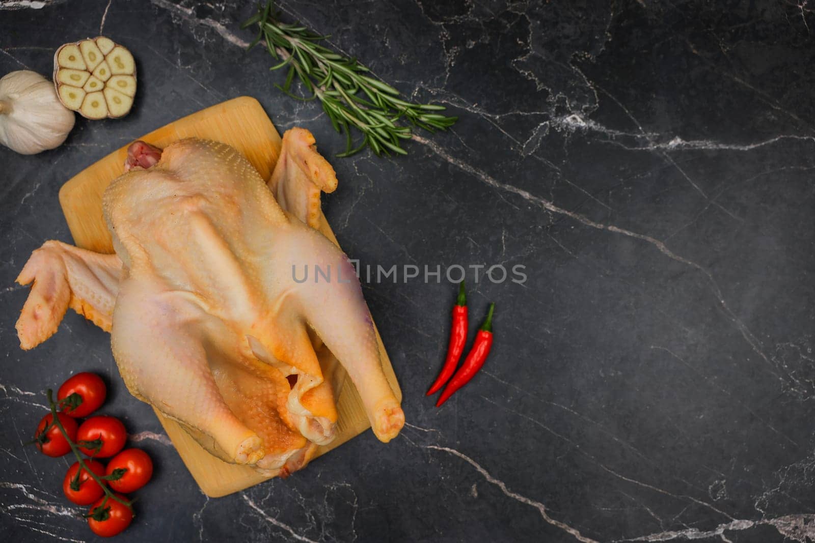 The butchered raw chicken lies on a wooden cutting board against a background of black marble, surrounded by tomatoes, red hot peppers, rosemary and garlic. The concept of cooking, diet menu, ingredients. High quality photo