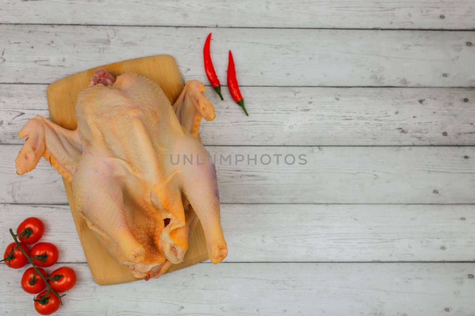 A close-up view from above of a butchered raw chicken lies on a wooden cutting board, with a bunch of tomatoes and red hot peppers on the sides against a white tree background with space for text. The concept of cooking, diet menu, ingredients. High quality photo