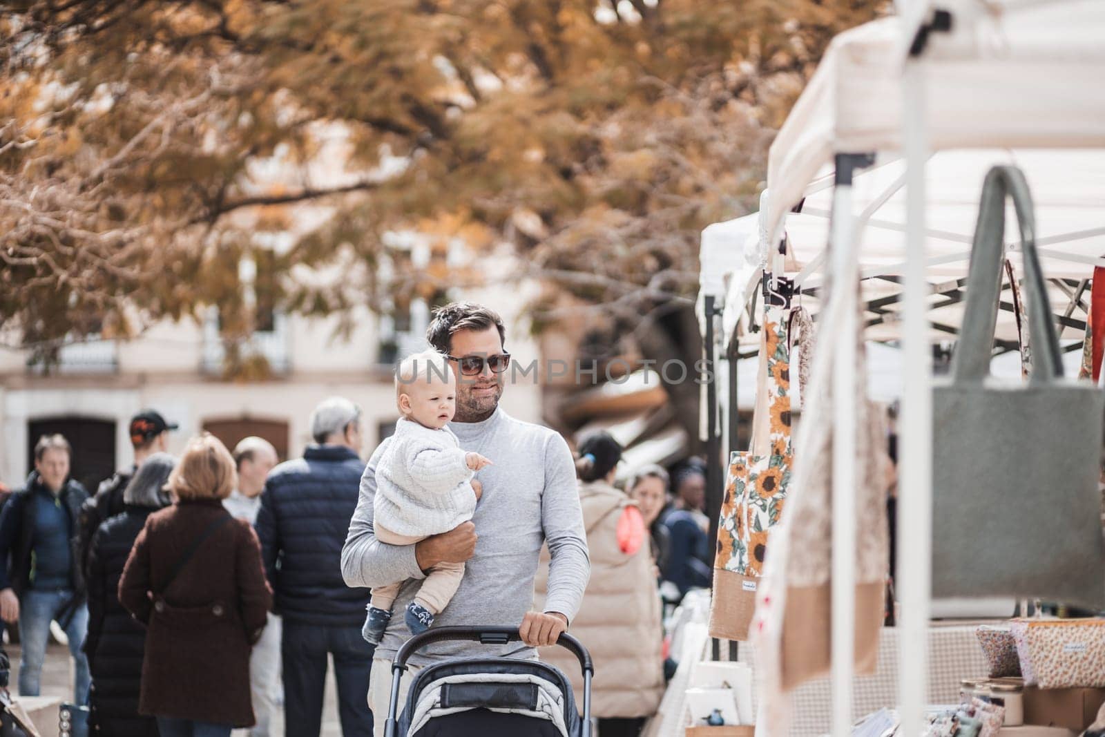 Father walking carrying his infant baby boy child and pushing stroller in crowd of people wisiting sunday flea market in Malaga, Spain by kasto