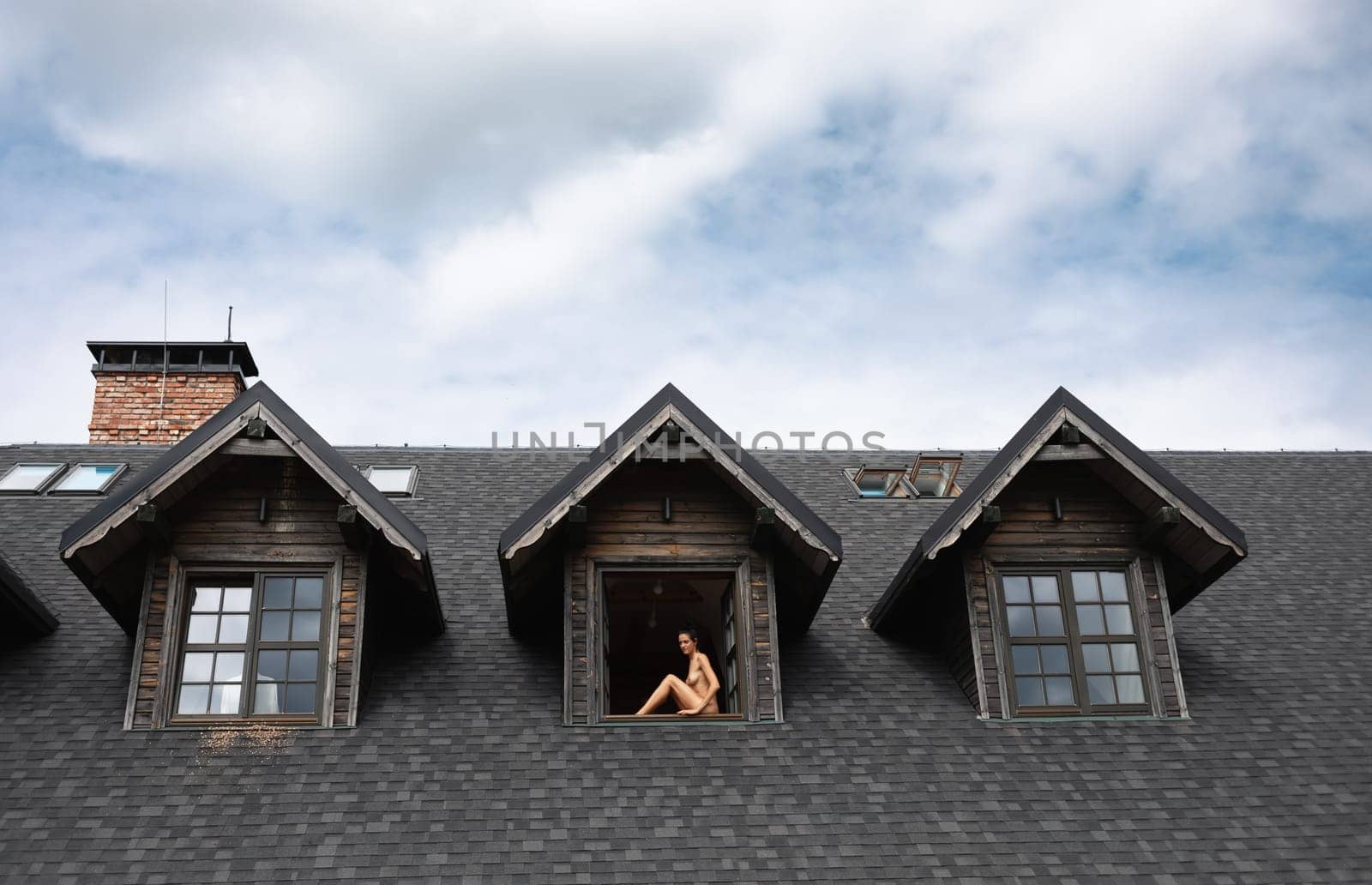 A young nude woman sits in the window opening of a country house enjoying a beautiful summer day