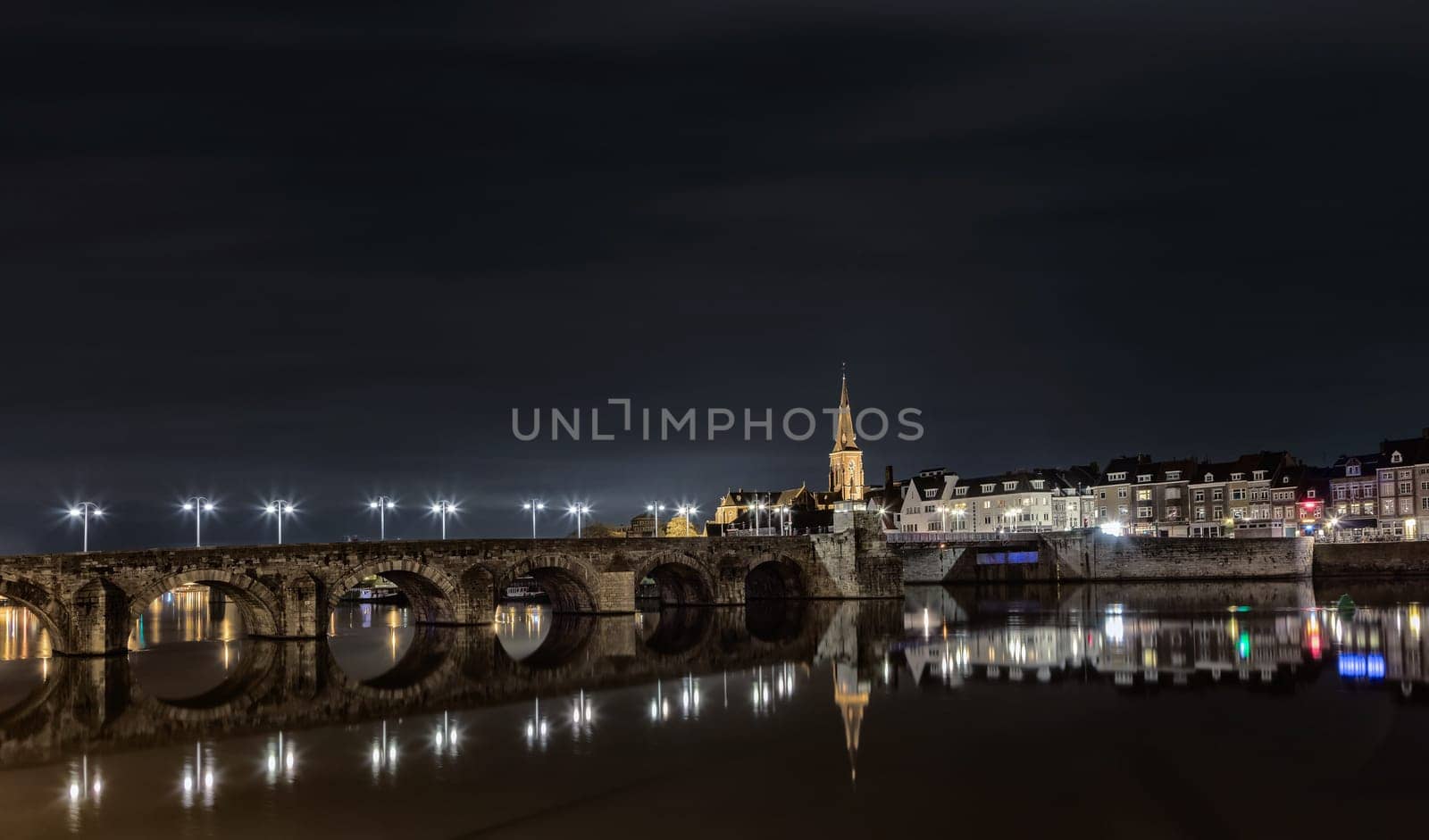 Riverside night scene in Maastricht, Netherlands with old bridge over river Maas and illuminated Saint Martin church.