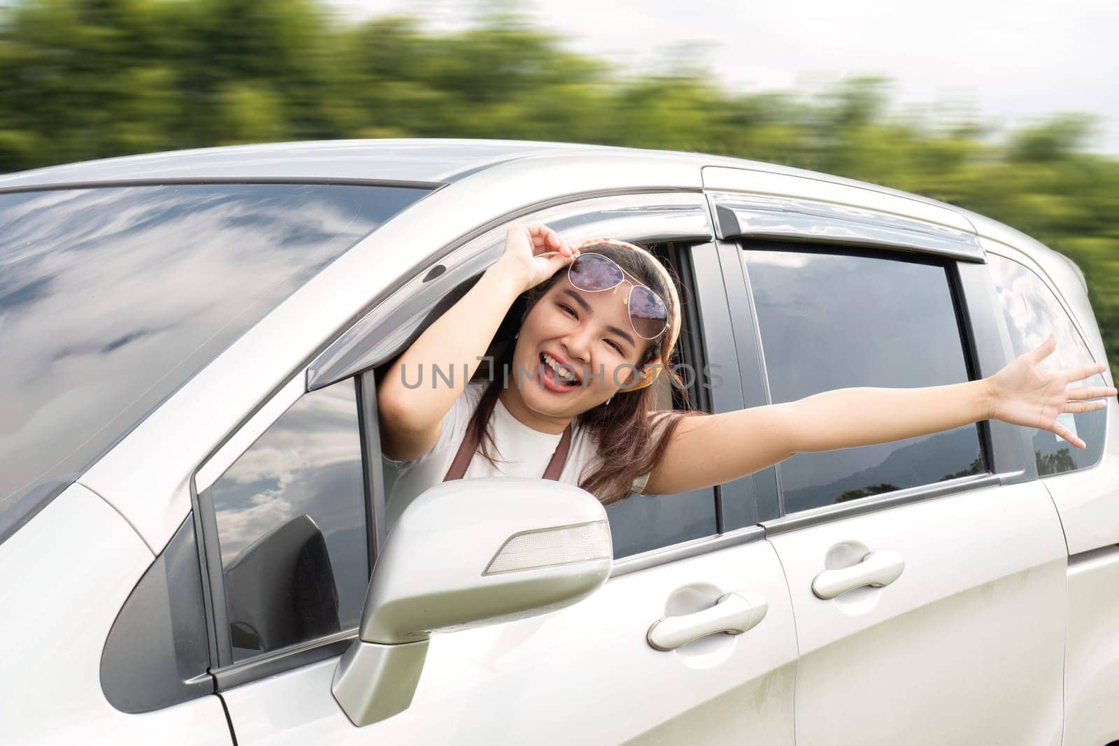 Happy young Asian woman enjoying a road trip and having fun waving at the air outside the car..