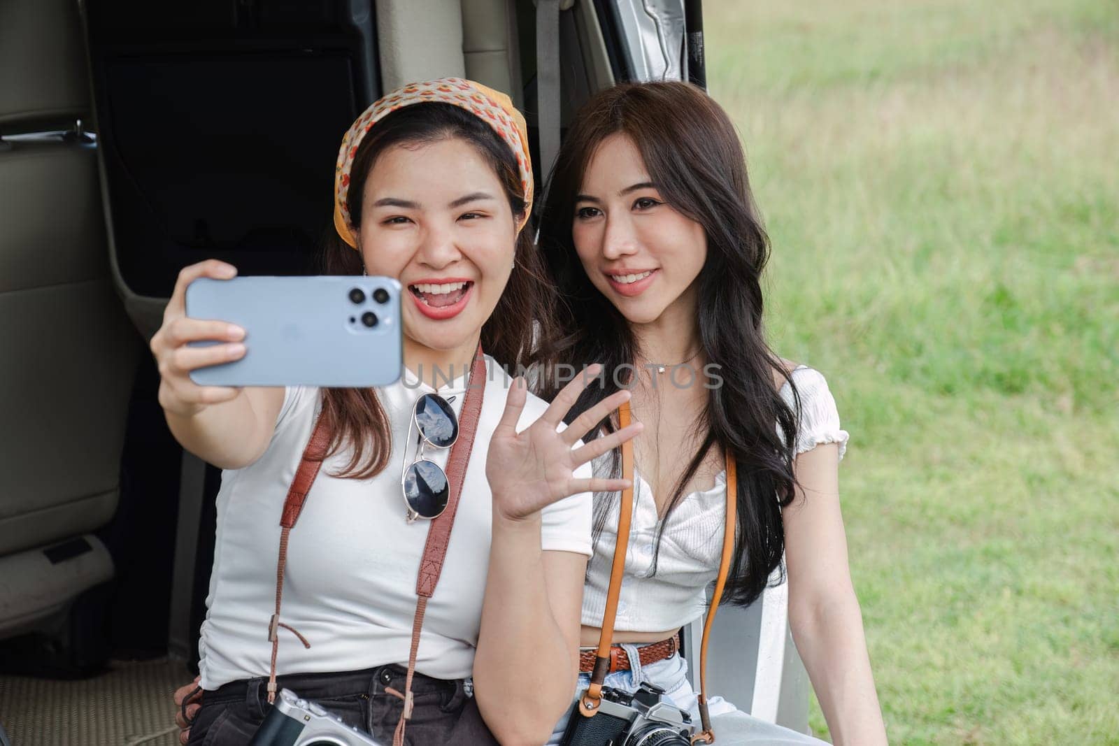 Two young women in white T-shirts and jeans sit back and relax, taking selfies together with their cell phones in the back of a car. by wichayada