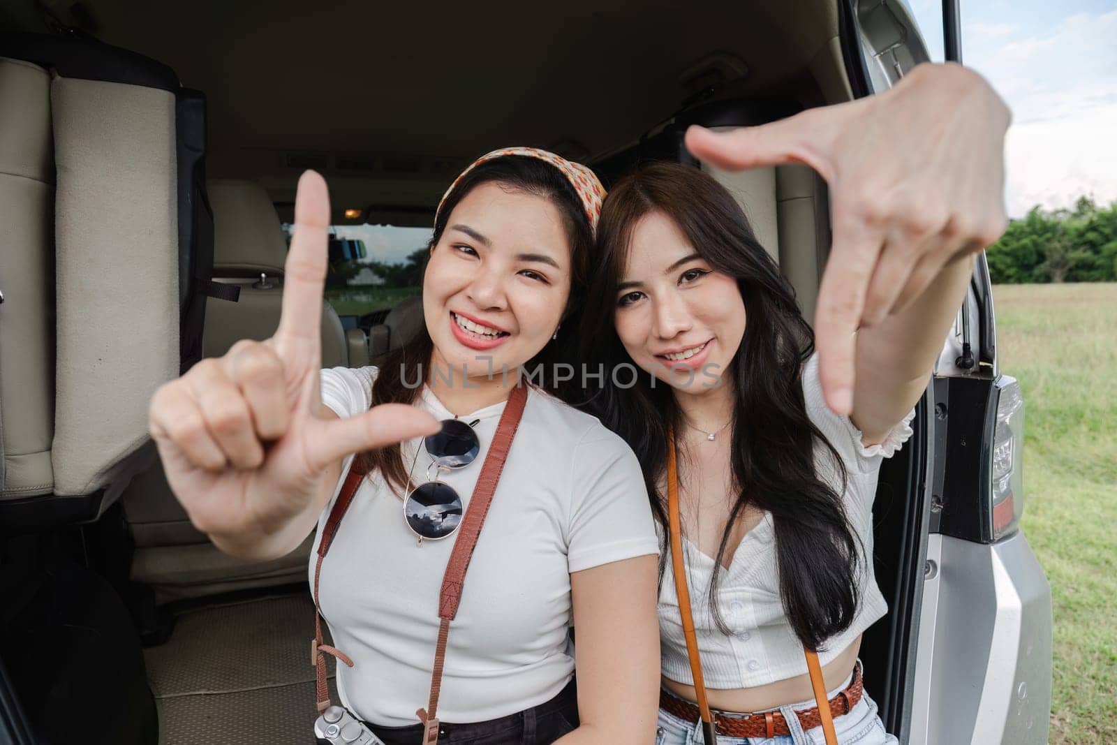 Two young Asian women in white shirts and jeans sit in the back of their new car, lounging on the field. holidays and traveling.