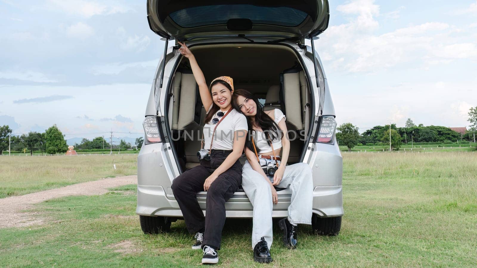 Two young Asian women in white shirts and jeans sit in the back of their new car, lounging on the field. holidays and traveling.