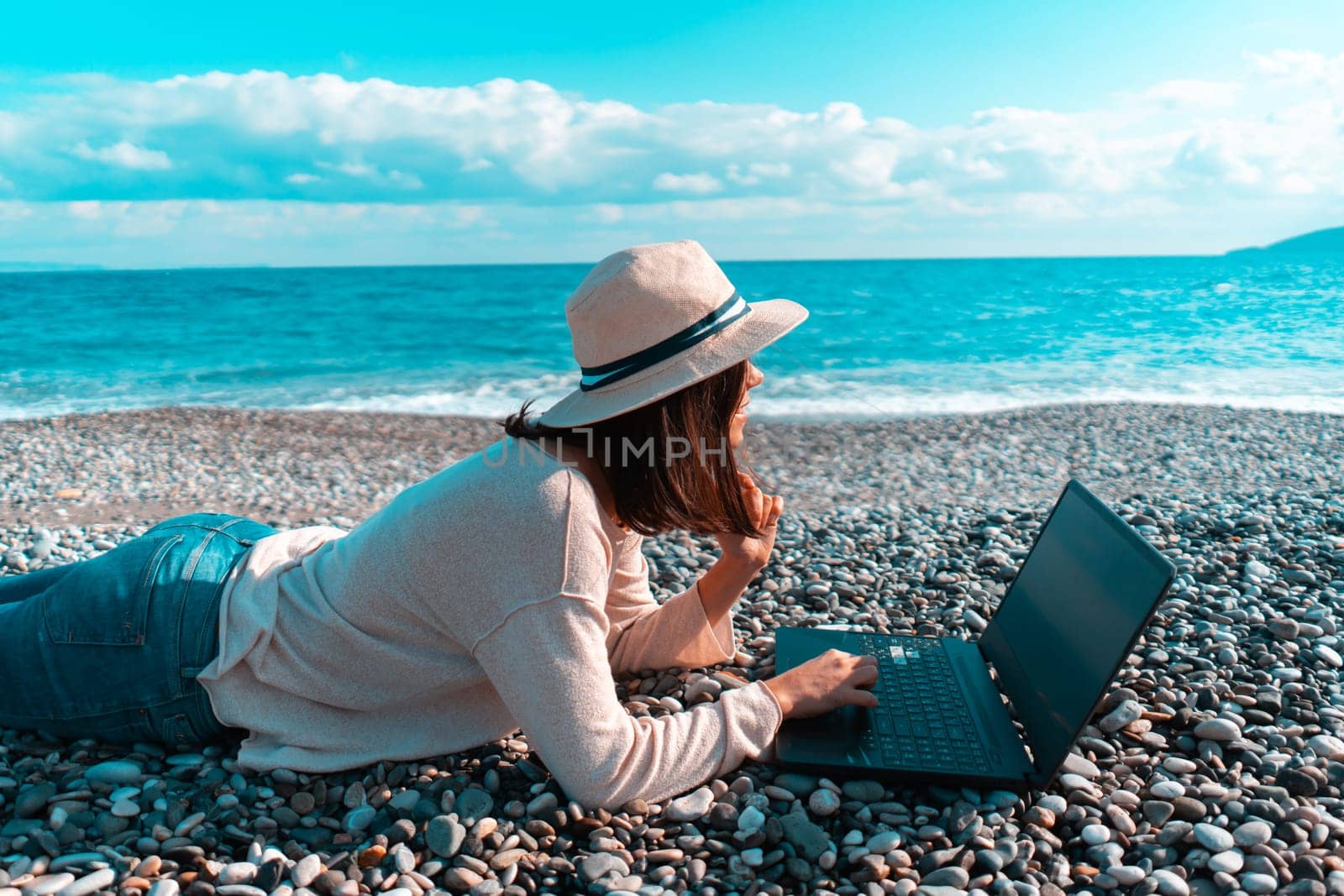 A young girl in a light hat and casual sweater lies on the beach by the sea with a laptop on a sunny day, works, studies, buys tickets during trip, a woman rests on vacation and types on the keyboard.