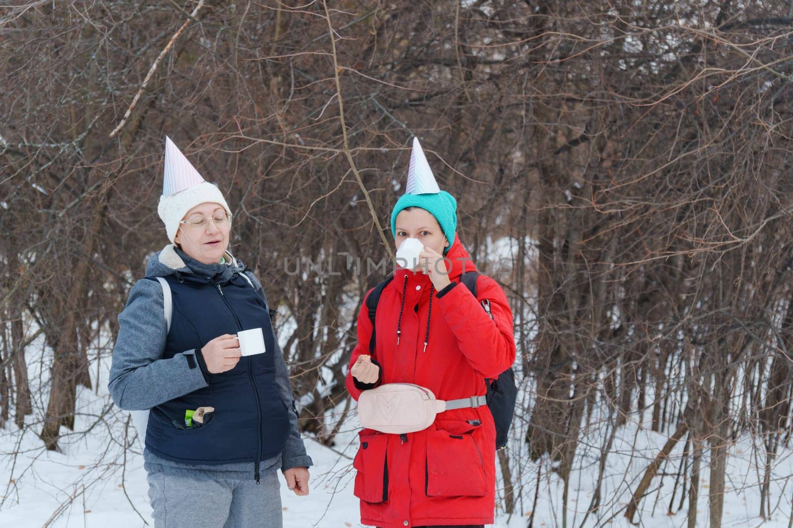 Two friends in festive party hats enjoy warm beverages while standing in a snowy area.