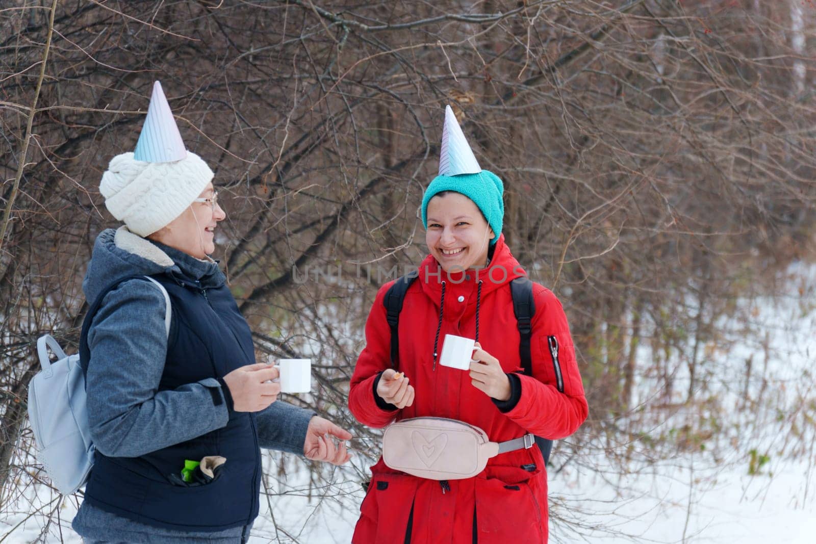 Two friends in festive party hats enjoy warm beverages while standing in a snowy area.