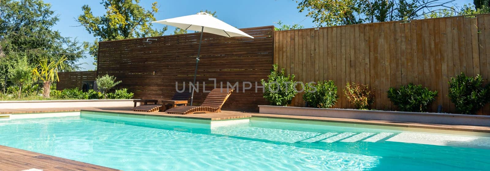 Relaxing backyard swimming pool with turquoise water, wooden deck, lounge chairs, umbrella, and green garden