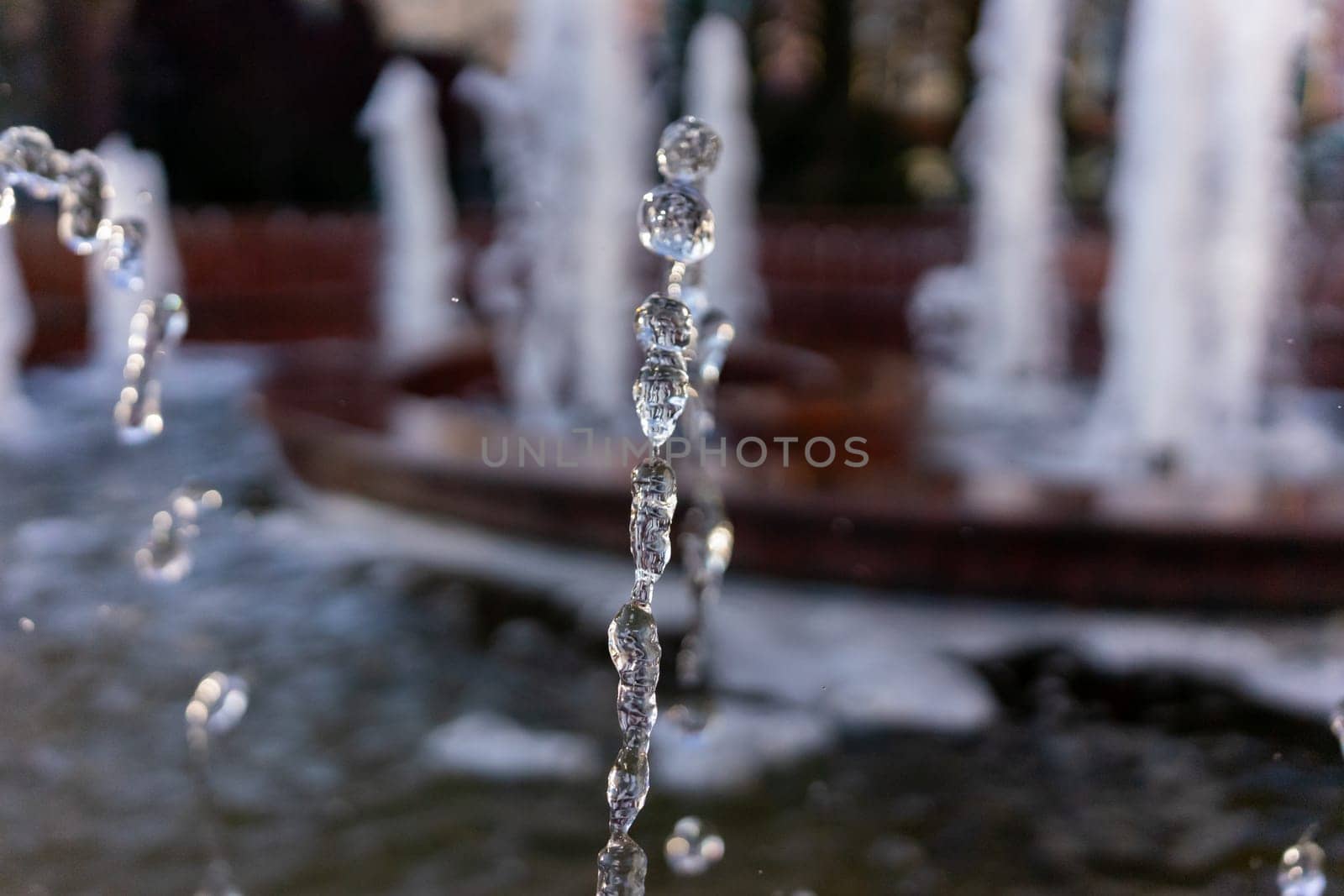 Water droplets emerge from a fountain, creating a mesmerizing display as they fall into the basin below. Sunlight sparkles on the water, enhancing the serene atmosphere of the park.
