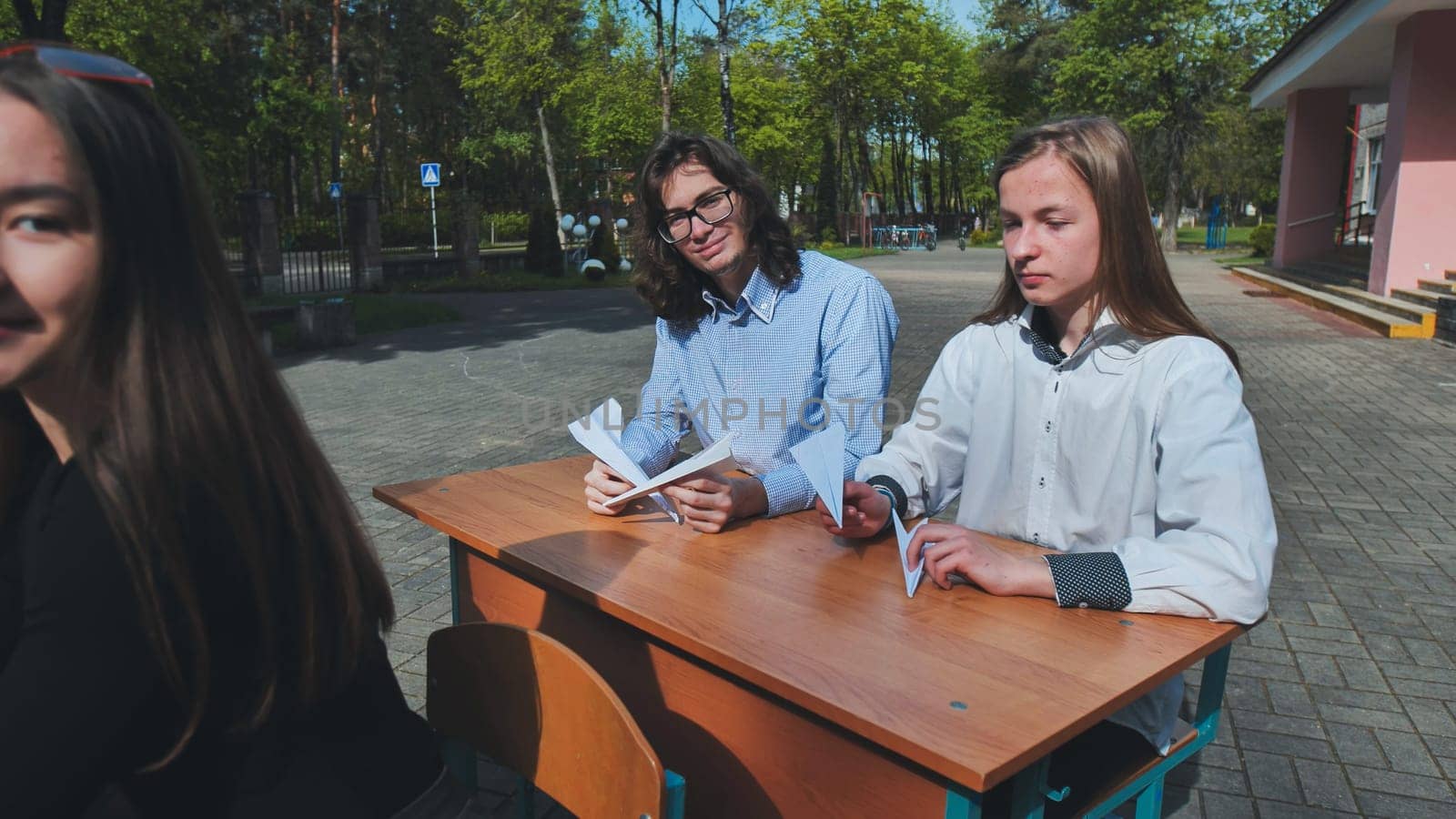 Two high school students are making paper airplanes at a desk outdoors on school grounds, enjoying a break