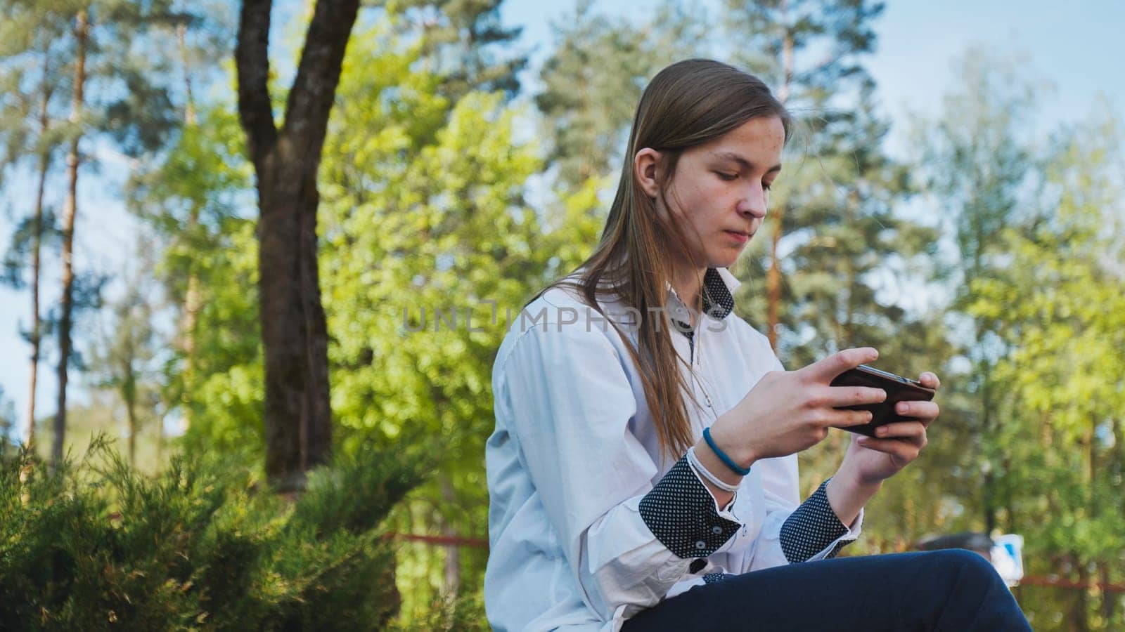 Teenager enjoying break time playing mobile games outdoors, concept of technology addiction and leisure during school day