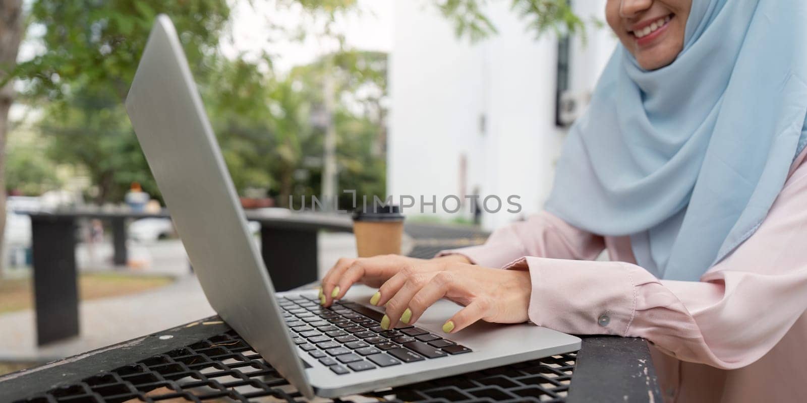 A focused Muslim woman in a hijab types on her laptop, with a coffee cup nearby, illustrating a modern approach to work and productivity in a serene outdoor setting.
