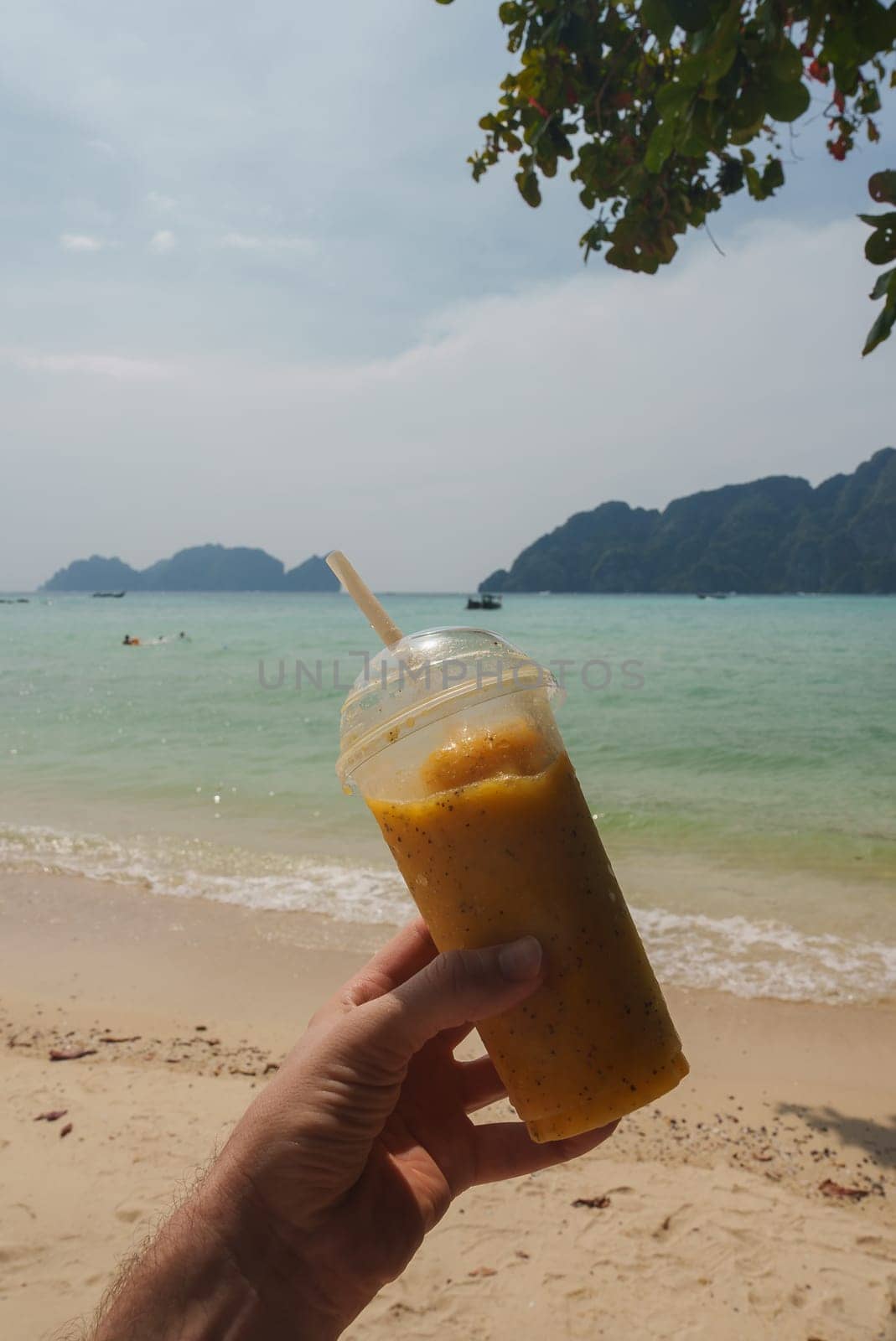 A vibrant yellow smoothie in a plastic cup held against a serene beach backdrop, with turquoise waters, lush islands, swimmers, and boats in Thailand.