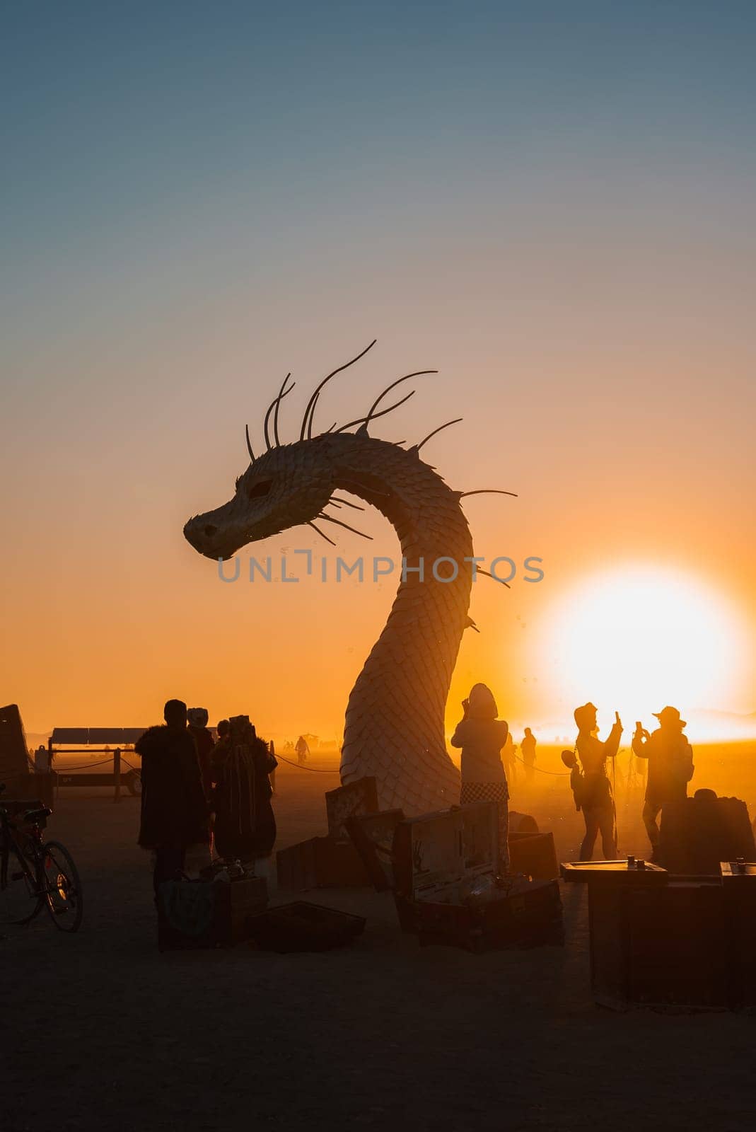 A large dragon like sculpture stands silhouetted against a vibrant sunset in a desert setting. Participants gather around, capturing photos and conversing.