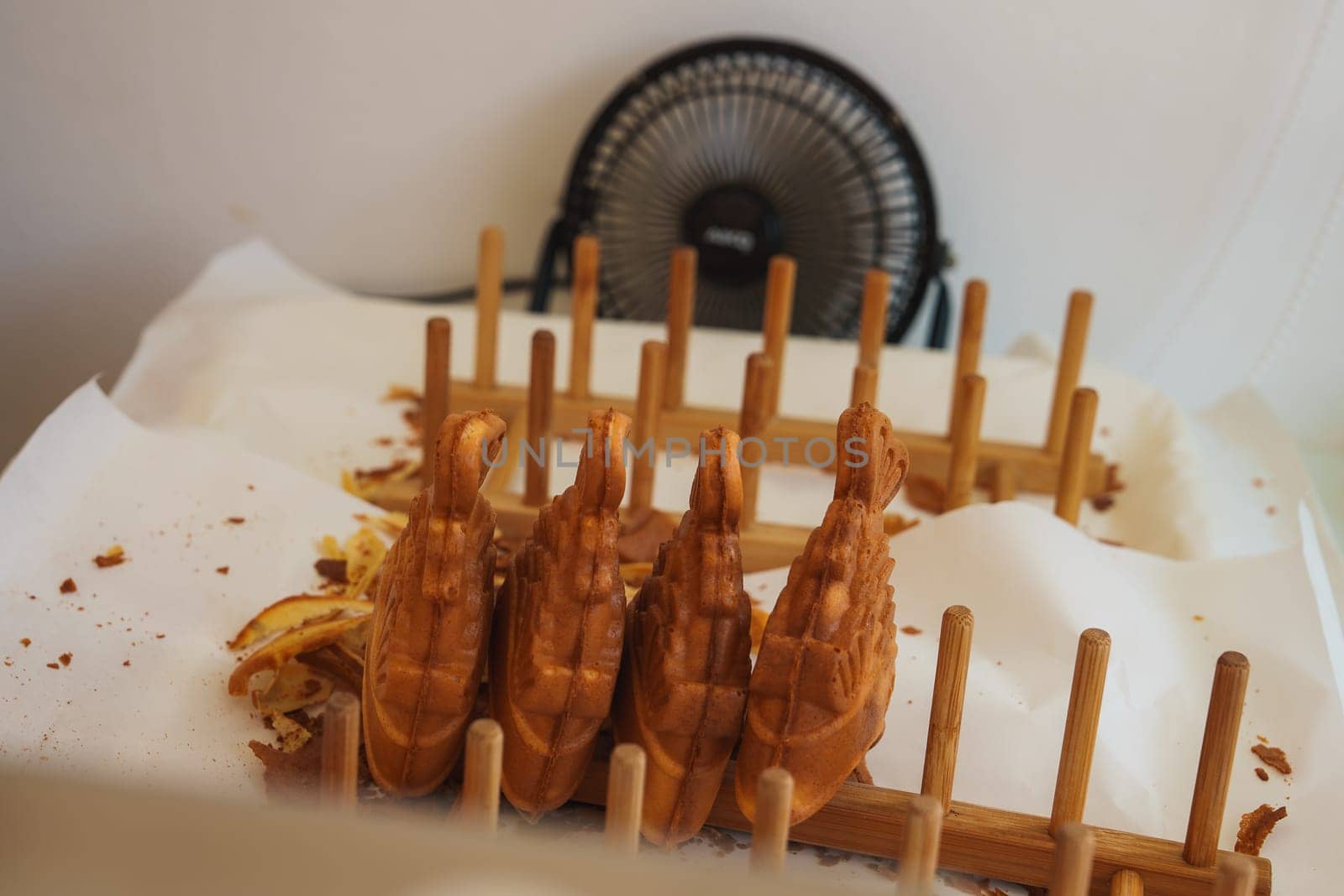 Golden brown swan shaped waffles displayed upright on a wooden rack with crumbs on white paper, a small black fan visible in the background.