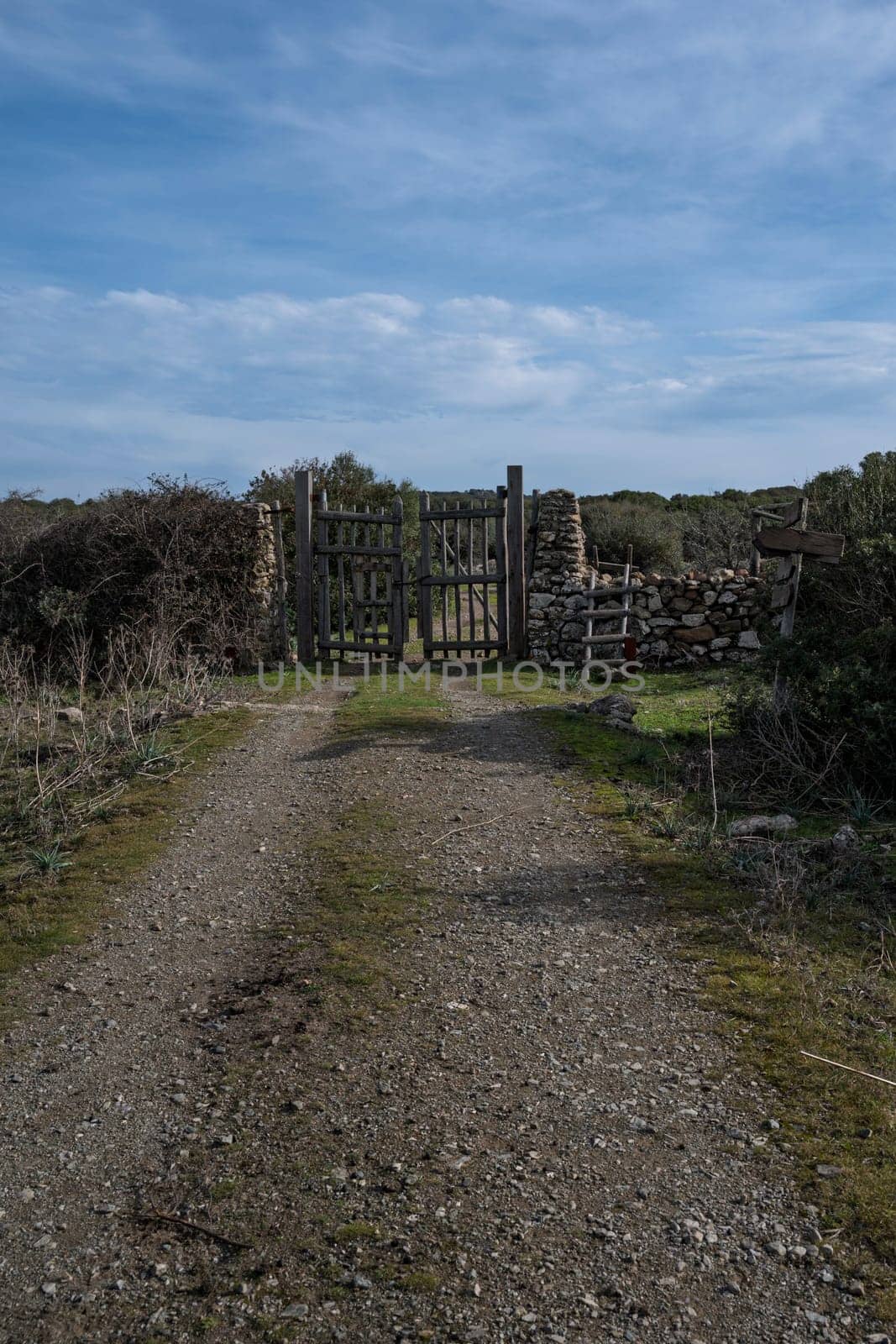 A rustic wooden gate with dry stone walls, located in a rural countryside with a dirt path and natural vegetation. The setting conveys tranquility and authenticity