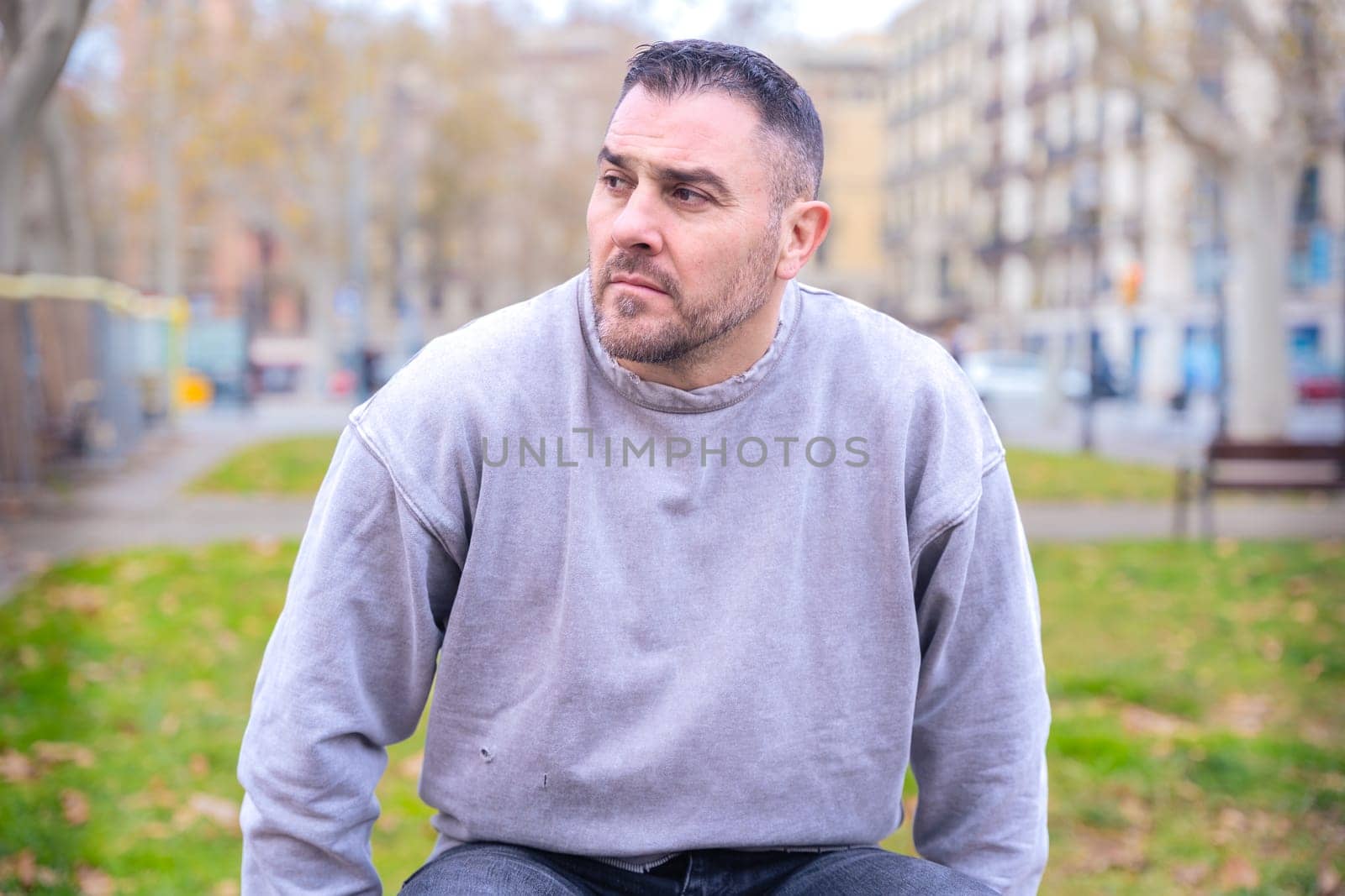 Man in a gray sweatshirt sitting on a bench in an urban park, looking thoughtful as he gazes into the distance. Background of trees, buildings, and an autumn city atmosphere.