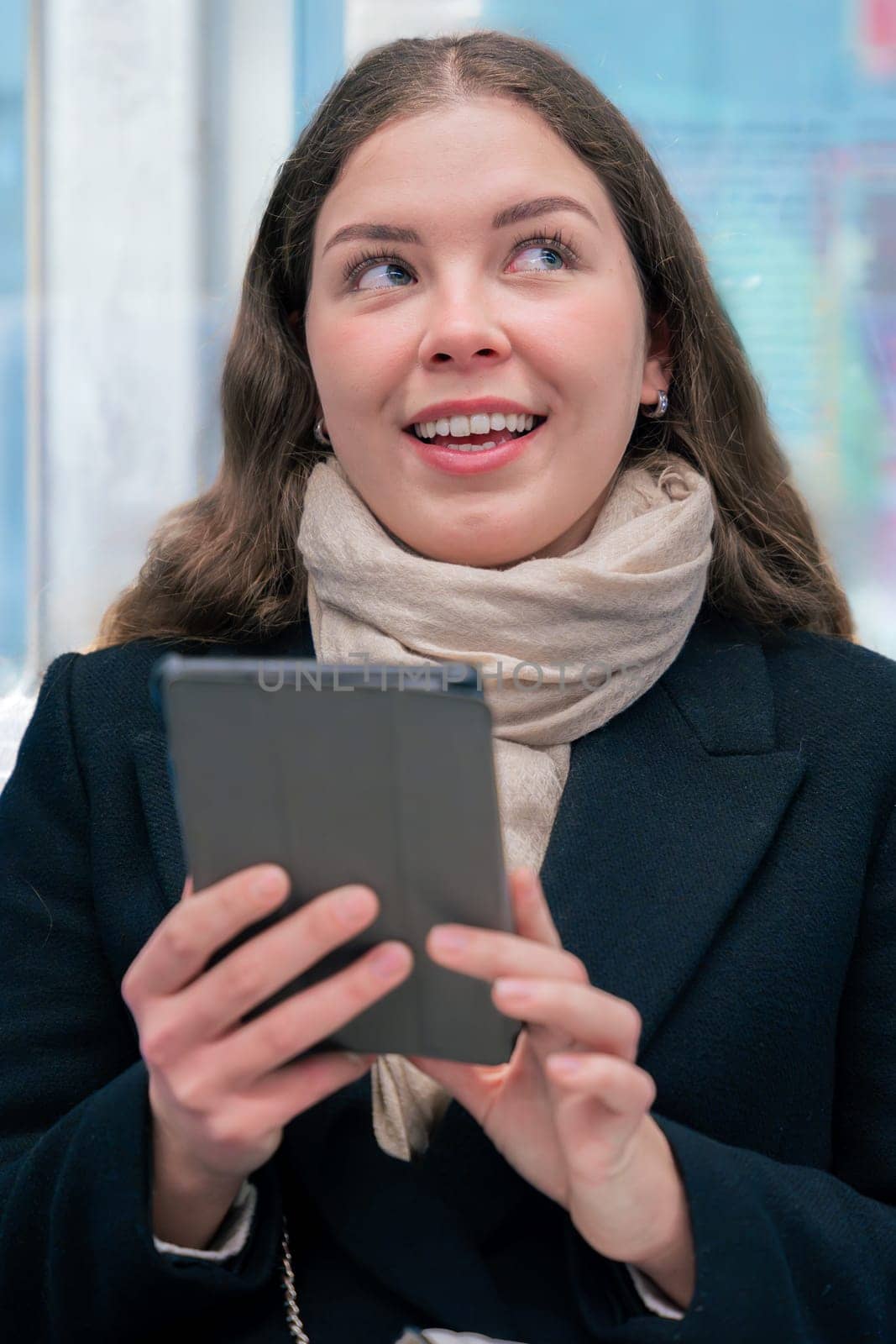 Focused young woman in a black coat and beige scarf sitting inside a modern subway train, reading on an e-reader. She enjoys a quiet moment during her daily commute.