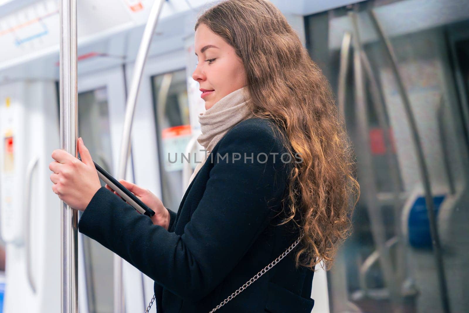 Stylish young woman in a black coat and beige scarf reading on an e-reader while standing inside a modern subway train. She balances while holding onto a pole during her commute.
