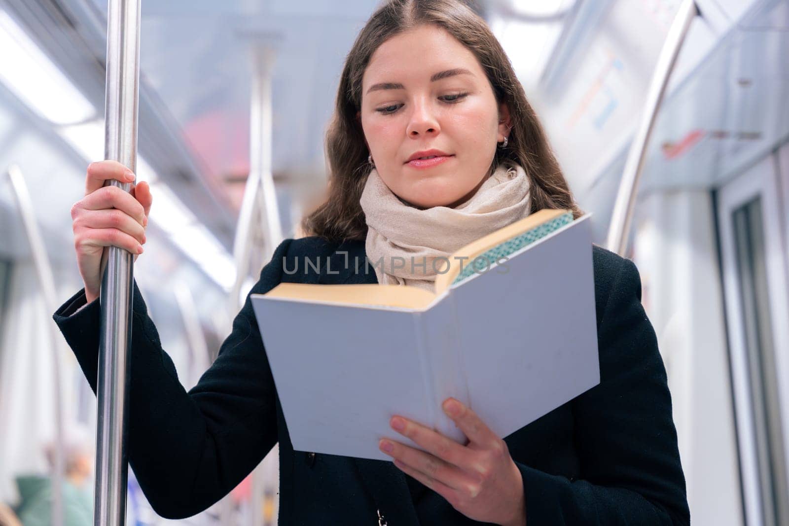 Focused young woman in a black coat and beige scarf standing inside a modern subway train, reading a book while holding onto a metal pole. She enjoys a quiet moment during her commute.