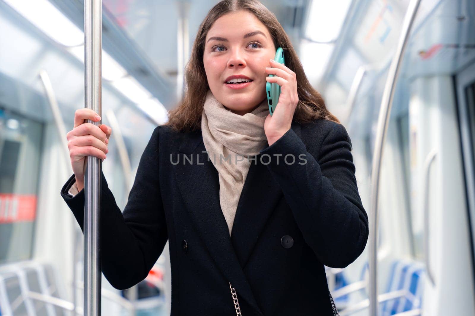 Cheerful young woman in a black coat and beige scarf talking on her smartphone while standing inside a modern subway train. She smiles brightly, holding onto a metal pole during her commute.