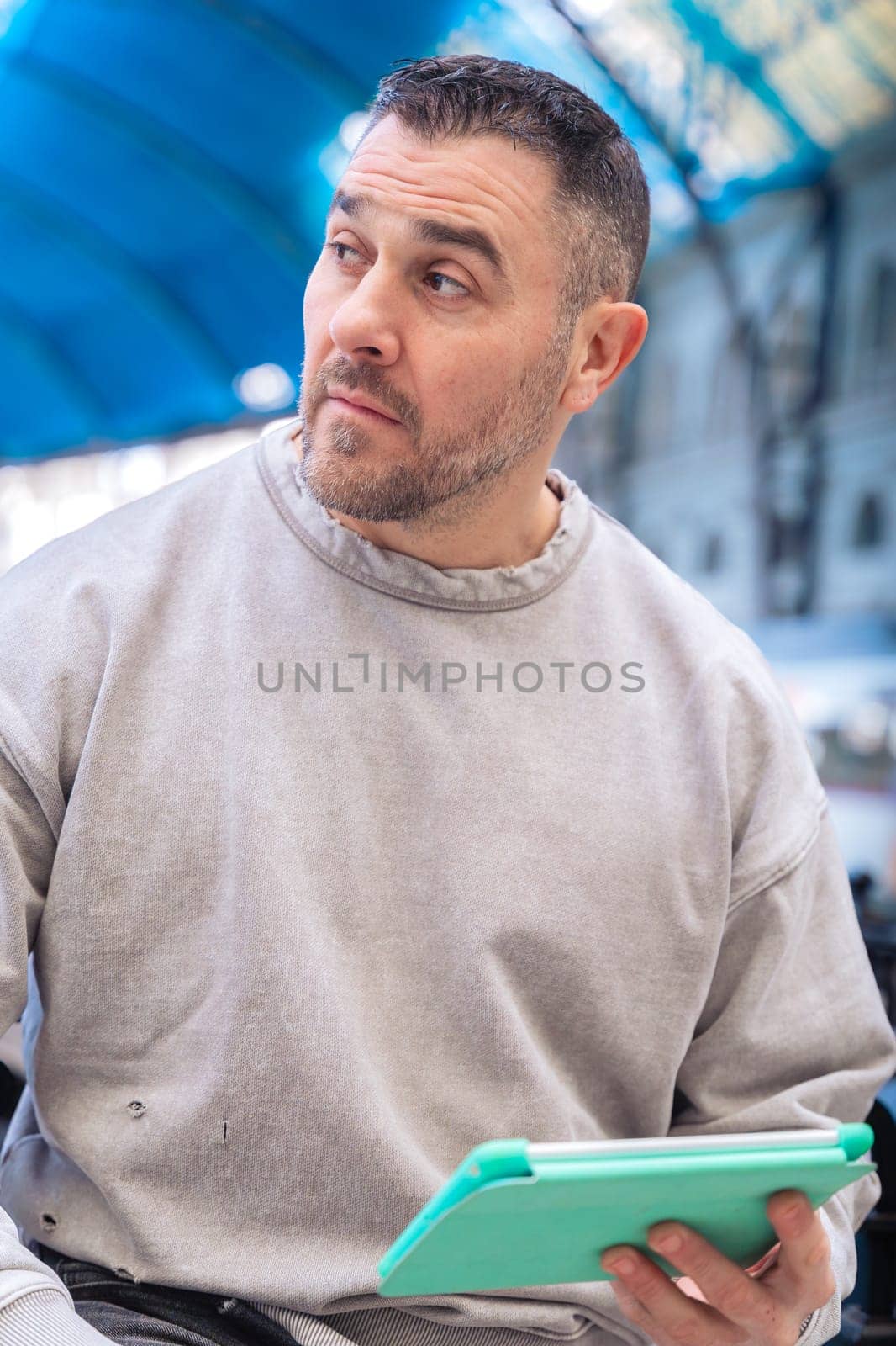Middle-aged man in a gray sweatshirt sitting at a train station, using a turquoise tablet. His serious expression and urban backdrop suggest work, travel, or digital connectivity.Vertical