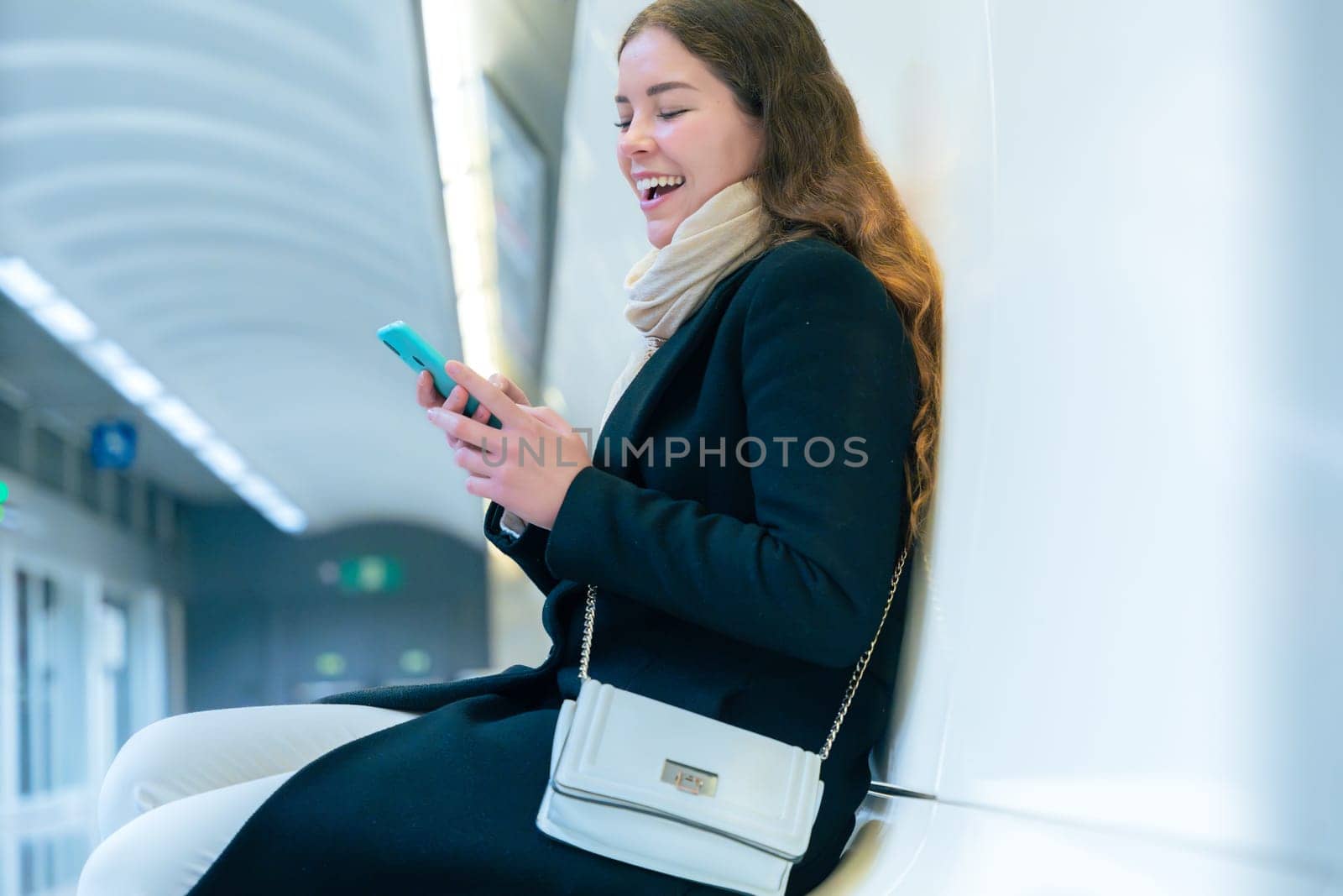 Stylish young woman in a black coat and scarf sitting in a modern subway station, smiling while using her smartphone. The bright and clean environment enhances the urban setting.