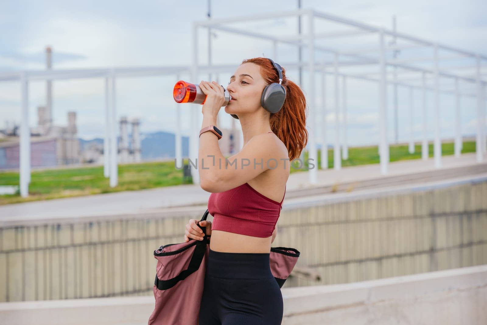 Athletic red-haired woman wearing sportswear and headphones, drinking water after exercise. She carries a gym bag and stands in a modern outdoor urban environment under a cloudy sky.