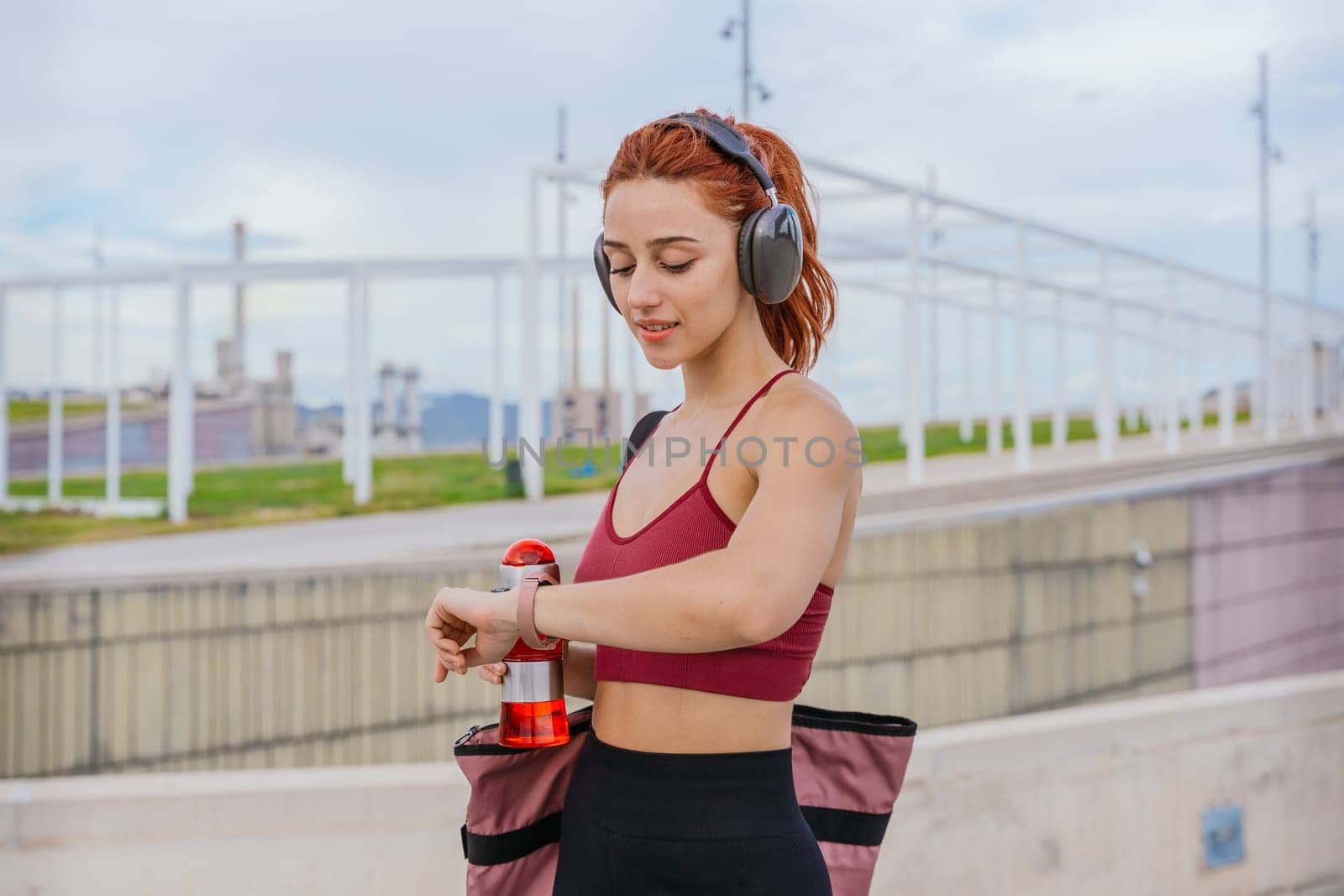 Fit young woman with red hair wearing sportswear and headphones, checking her smartwatch while holding a water bottle. She carries a gym bag and stands in a modern outdoor urban environment.