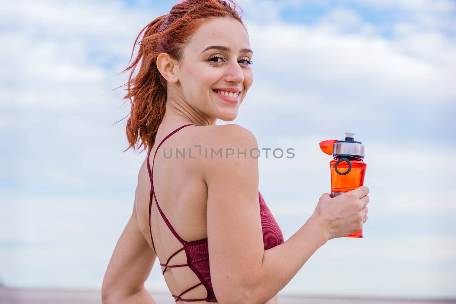 Fit young woman with red hair wearing a stylish sports bra, smiling while holding a water bottle. She stands outdoors under a bright blue sky, radiating positivity and energy after a workout.